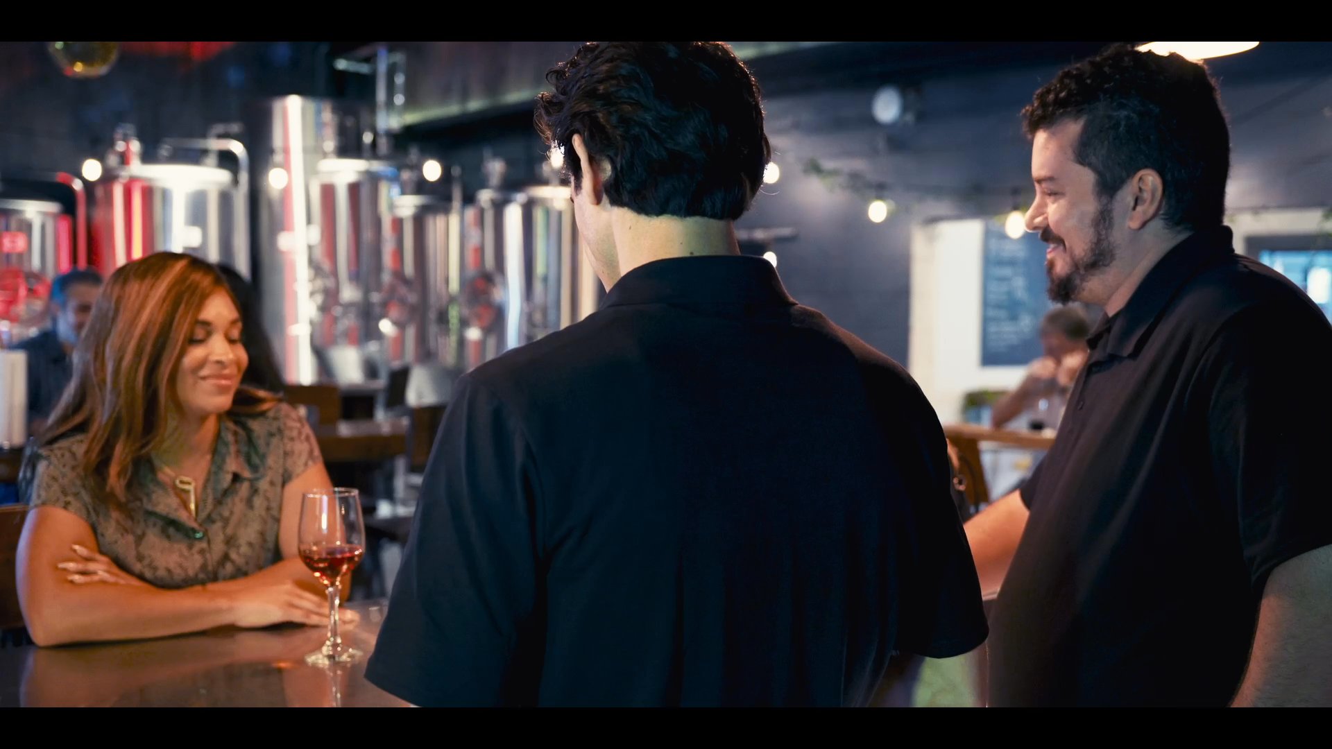 A woman engages in conversation with two brewery staff members while seated at the bar. The staff attend to their duties while maintaining a professional rapport with the patron.