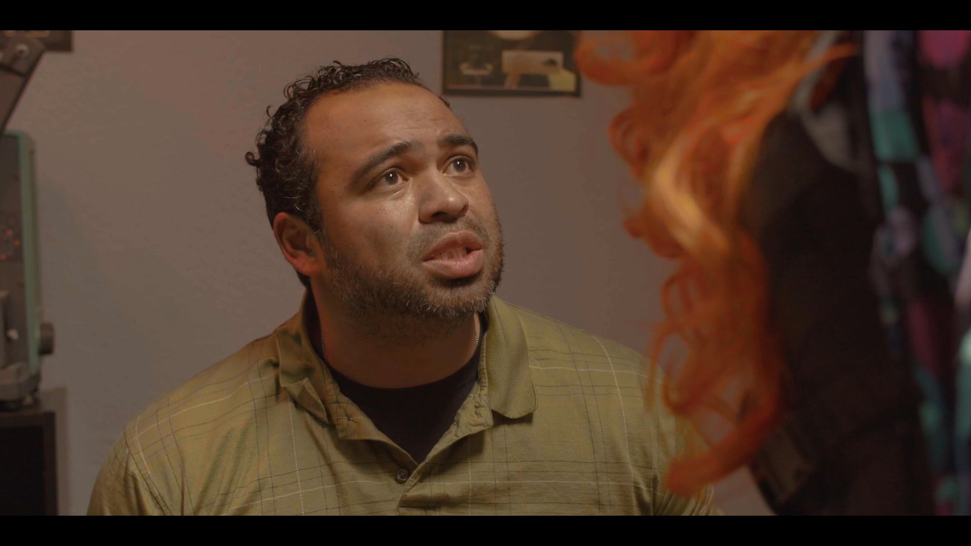 A person in drag engages in a tense, emotional conversation with an associate while a woman sits nearby crying. The scene captures a moment of interpersonal conflict and heightened vulnerability within an indoor setting.