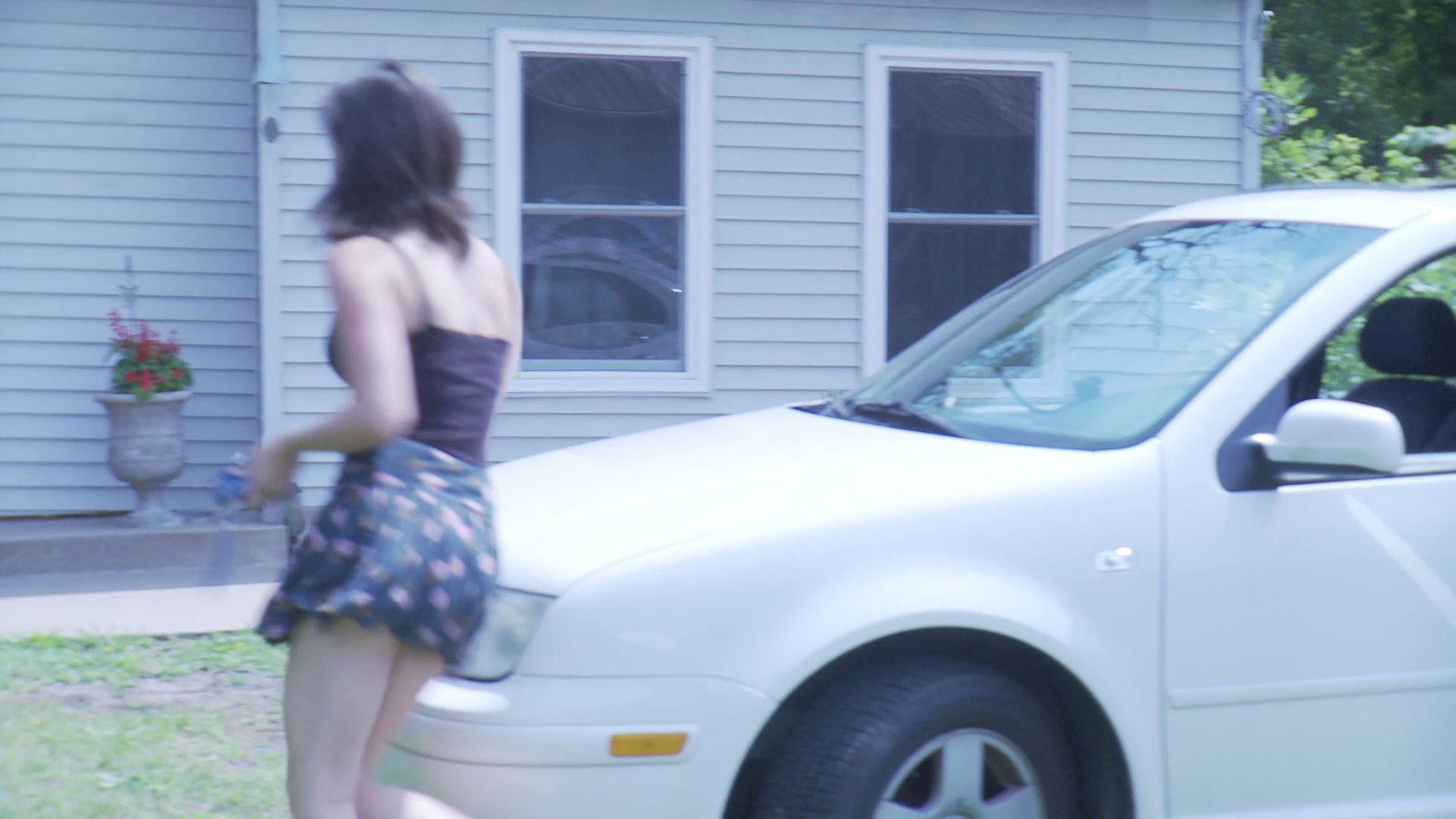 A young woman parks her car in a suburban driveway and walks toward the front door of a home. Upon entering the house, she finds an older man sitting inside.