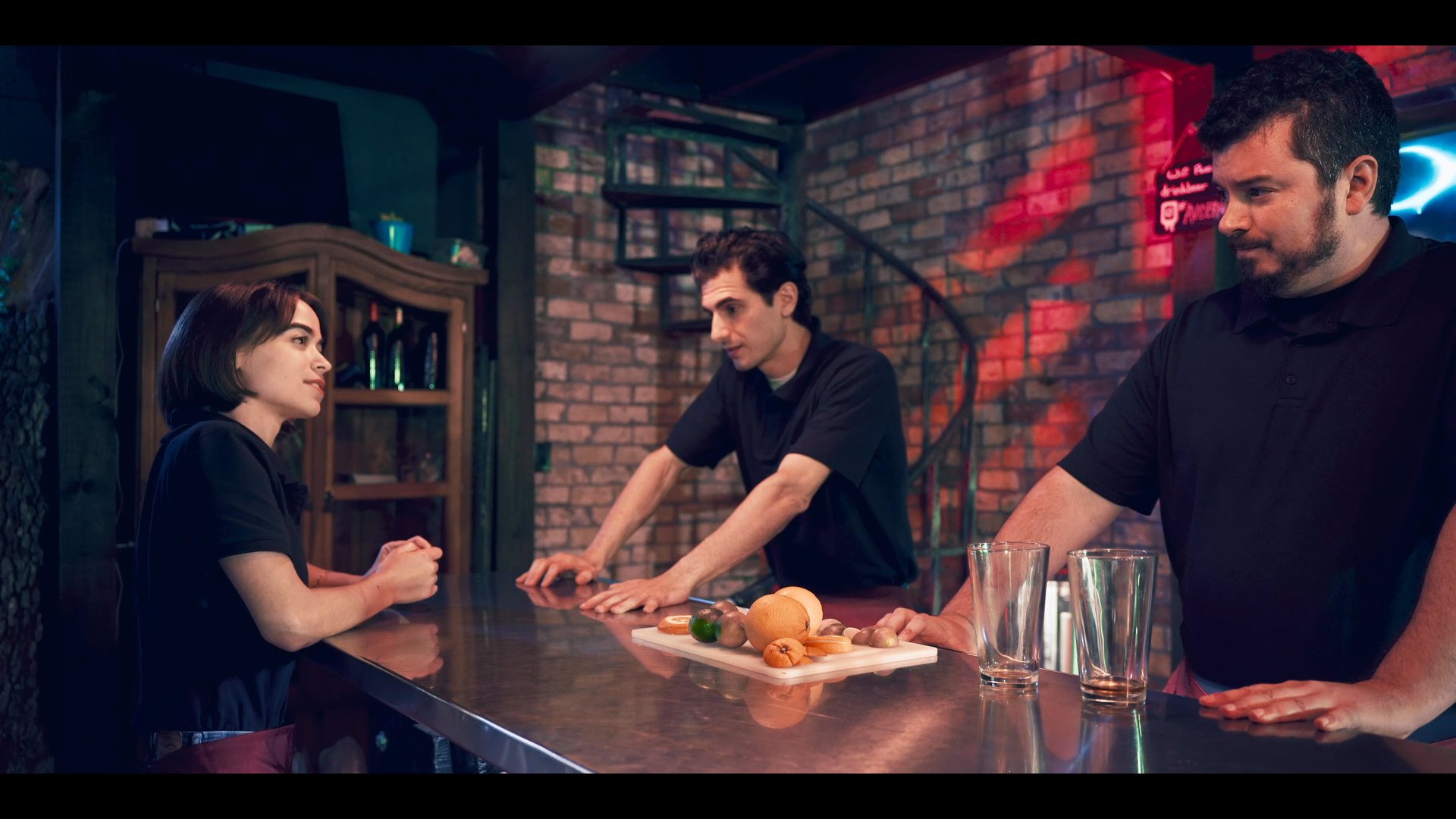 A bartender engages in a professional discussion with customers at a bar counter while another individual sits nearby holding a beverage.