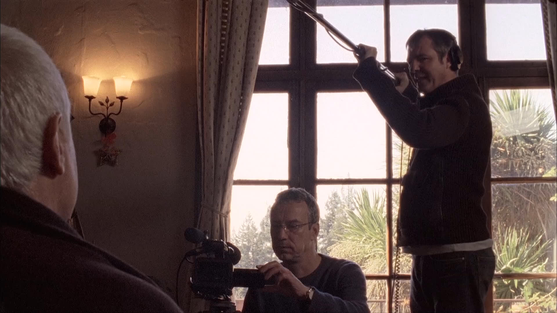 A subject discusses filmmaking techniques during an interview while a camera operator adjusts a lighting boom in the background.