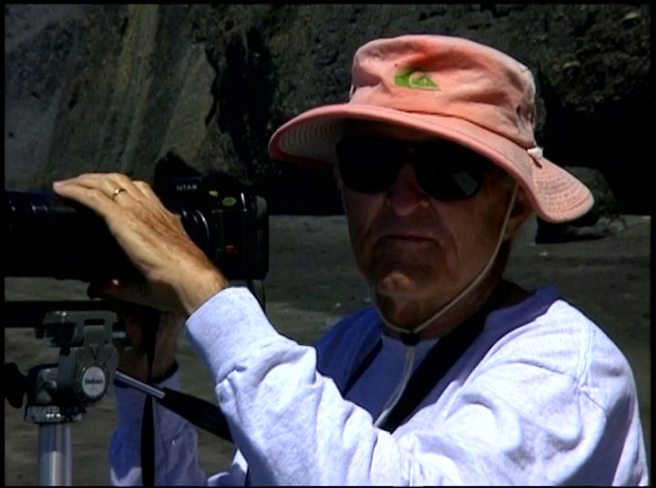 A photographer documents surfers on the ocean from the shoreline using a professional camera and telephoto lens. The scene transitions between dynamic action shots of riders on waves and the focused work of the photographer on the beach.