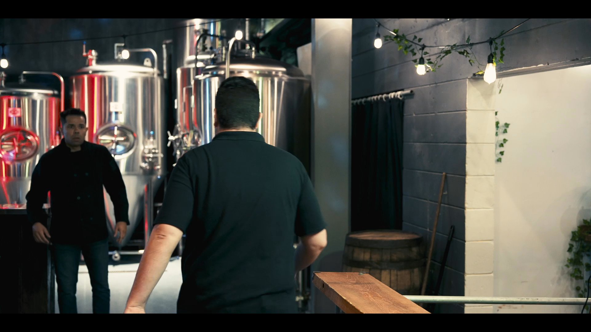 Two individuals conduct a guided walkthrough of a professional brewing facility, concluding as one exits the production area into an exterior courtyard.