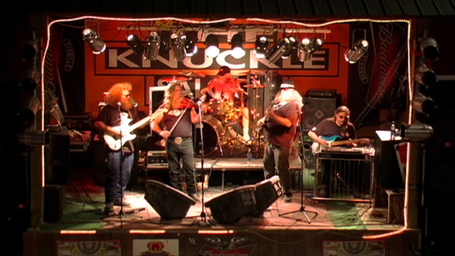 A long-haired male musician alternates between playing an electric guitar and a violin during a live stage performance. He performs alongside a band in a dimly lit venue.
