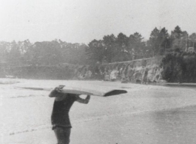 A person in vintage swimwear walks along a beach carrying a wooden surfboard on their head. The scene transitions to a panoramic view of a busy coastal area near a pier.