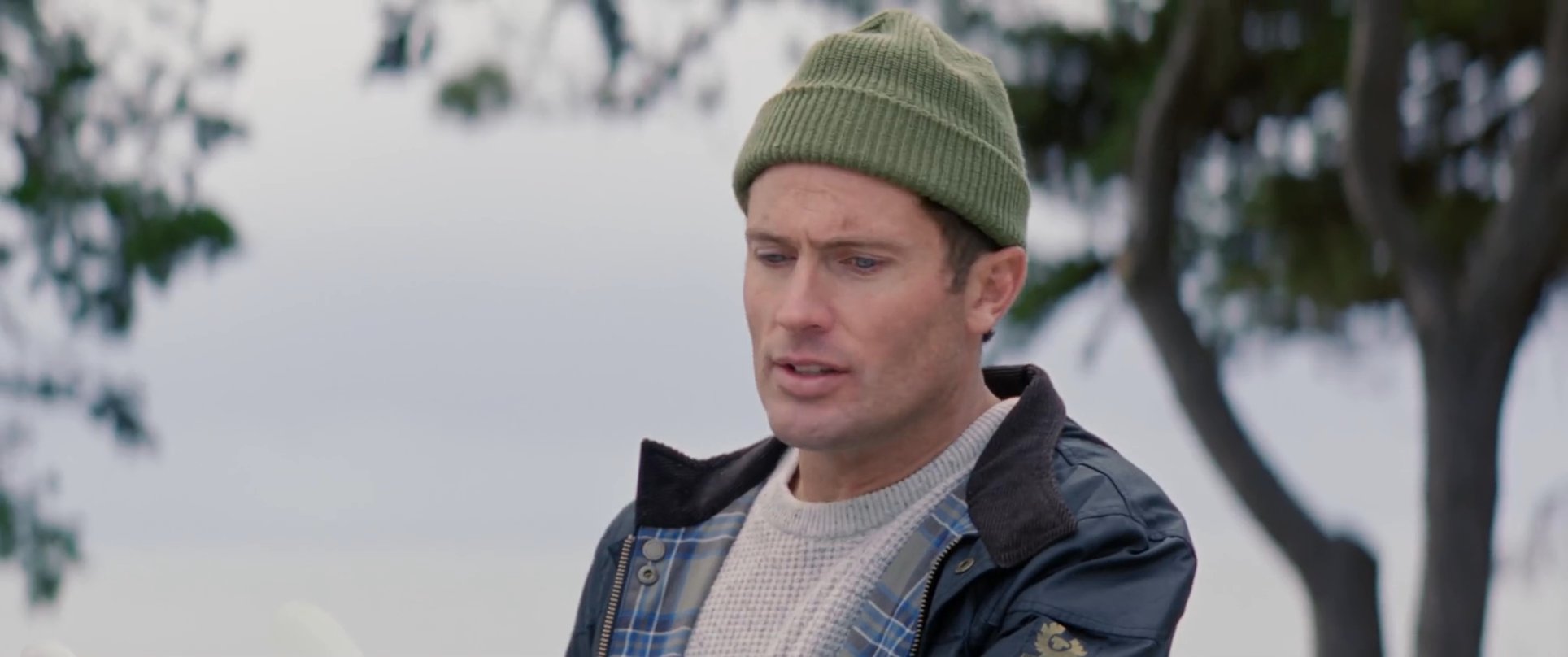 Two men wearing winter-appropriate coastal attire engage in a serious discussion while standing near their surfboards at a beach. A third individual observes the exchange in the background.