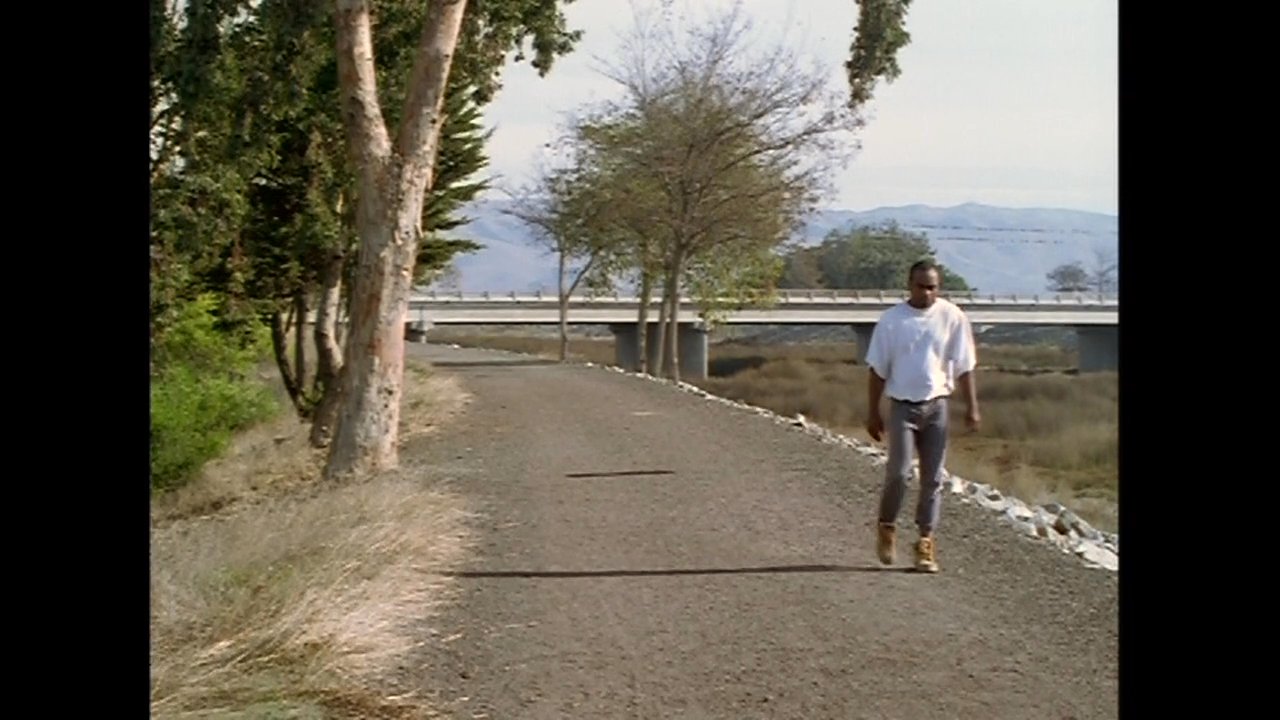 A man in a white t-shirt and jeans walks along a gravel trail bordered by trees and expansive hilly terrain. He moves with a steady, rhythmic pace across the open, natural landscape.