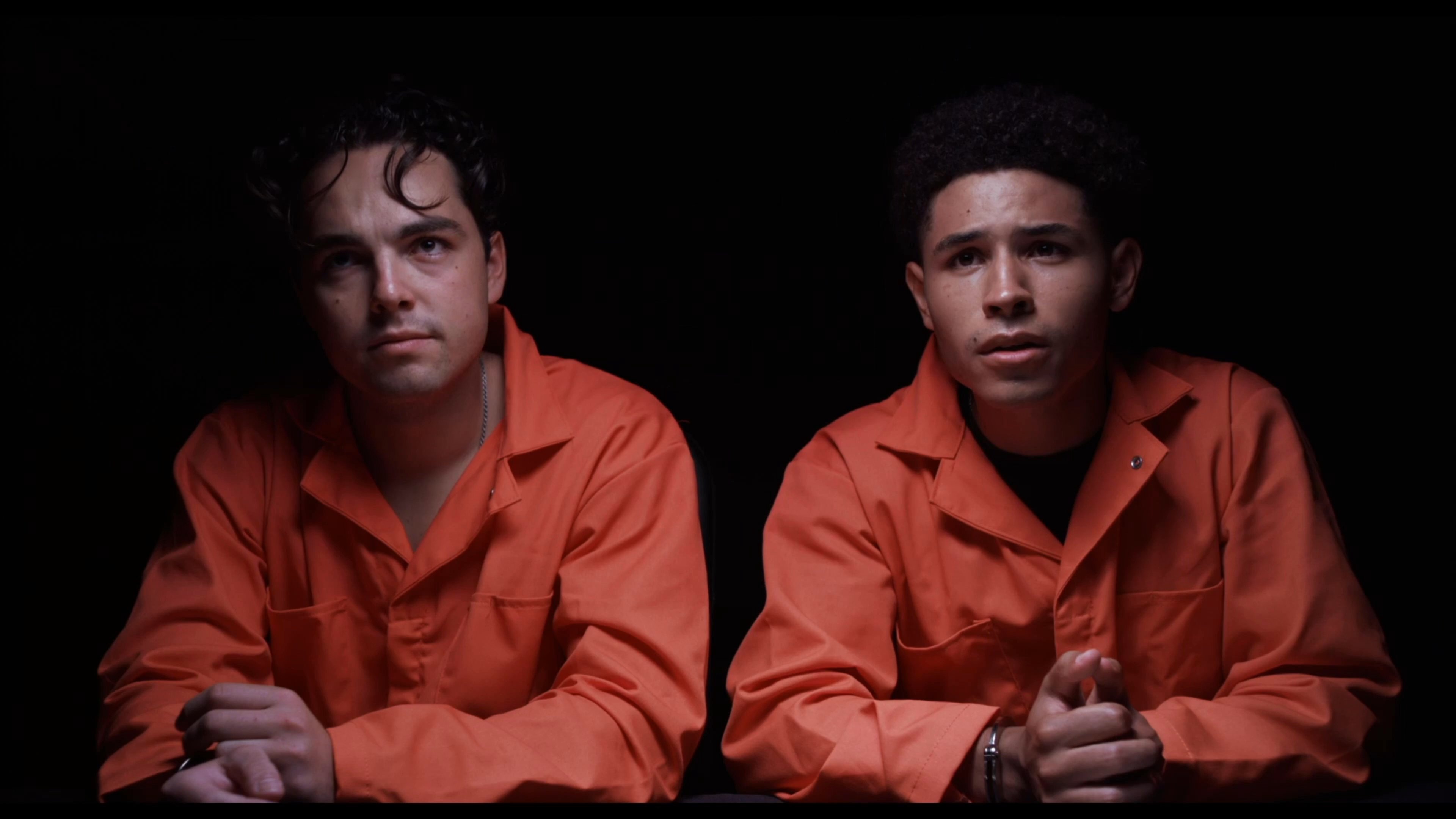A DEA agent interviews two suspects in orange jumpsuits before a third individual wearing a provocative t-shirt enters the room.