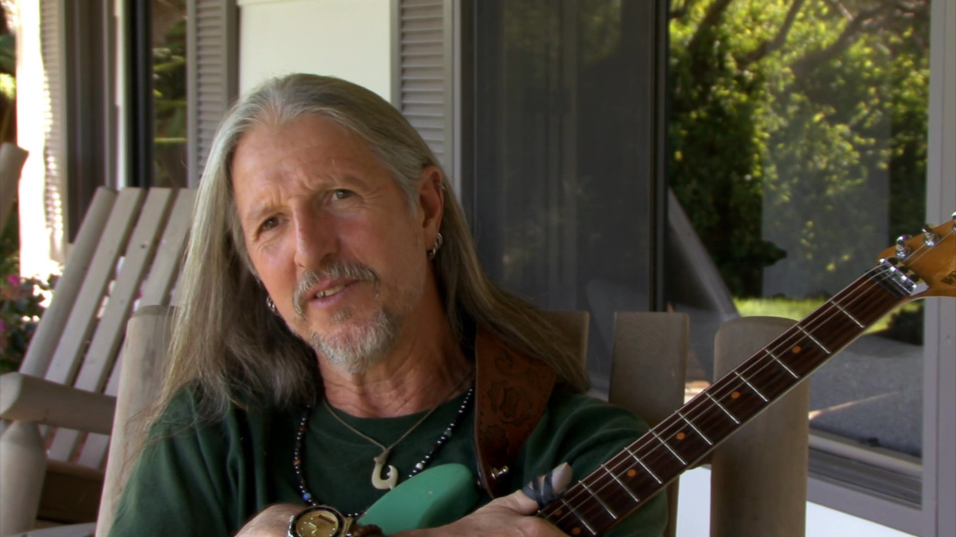 A long-haired musician sits with an acoustic guitar while engaging in a thoughtful conversation in an indoor setting. He speaks expressively, sharing insights while holding his instrument.