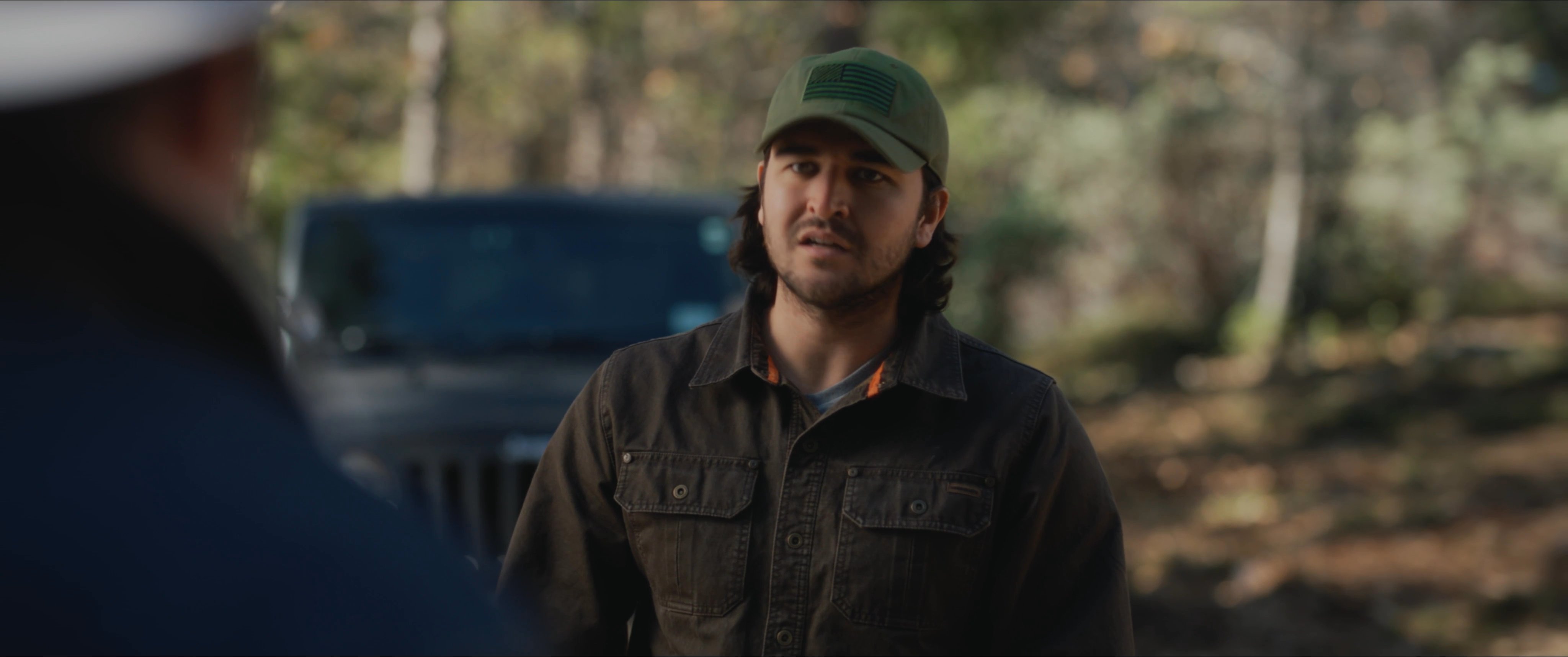 Two men have a serious discussion in a wooded area while one smokes a cigar, creating a tense atmosphere.