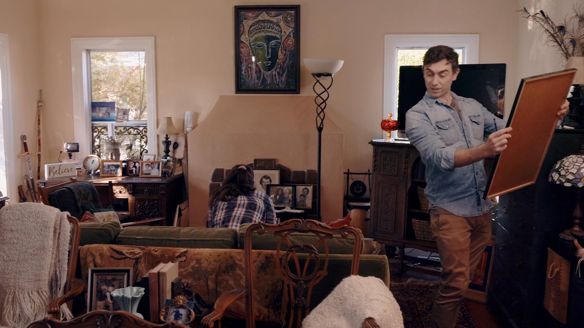 A man installs a framed Grateful Dead poster on a wall in a living room while a woman organizes items nearby. The pair works together to personalize their domestic space with decorative artwork.