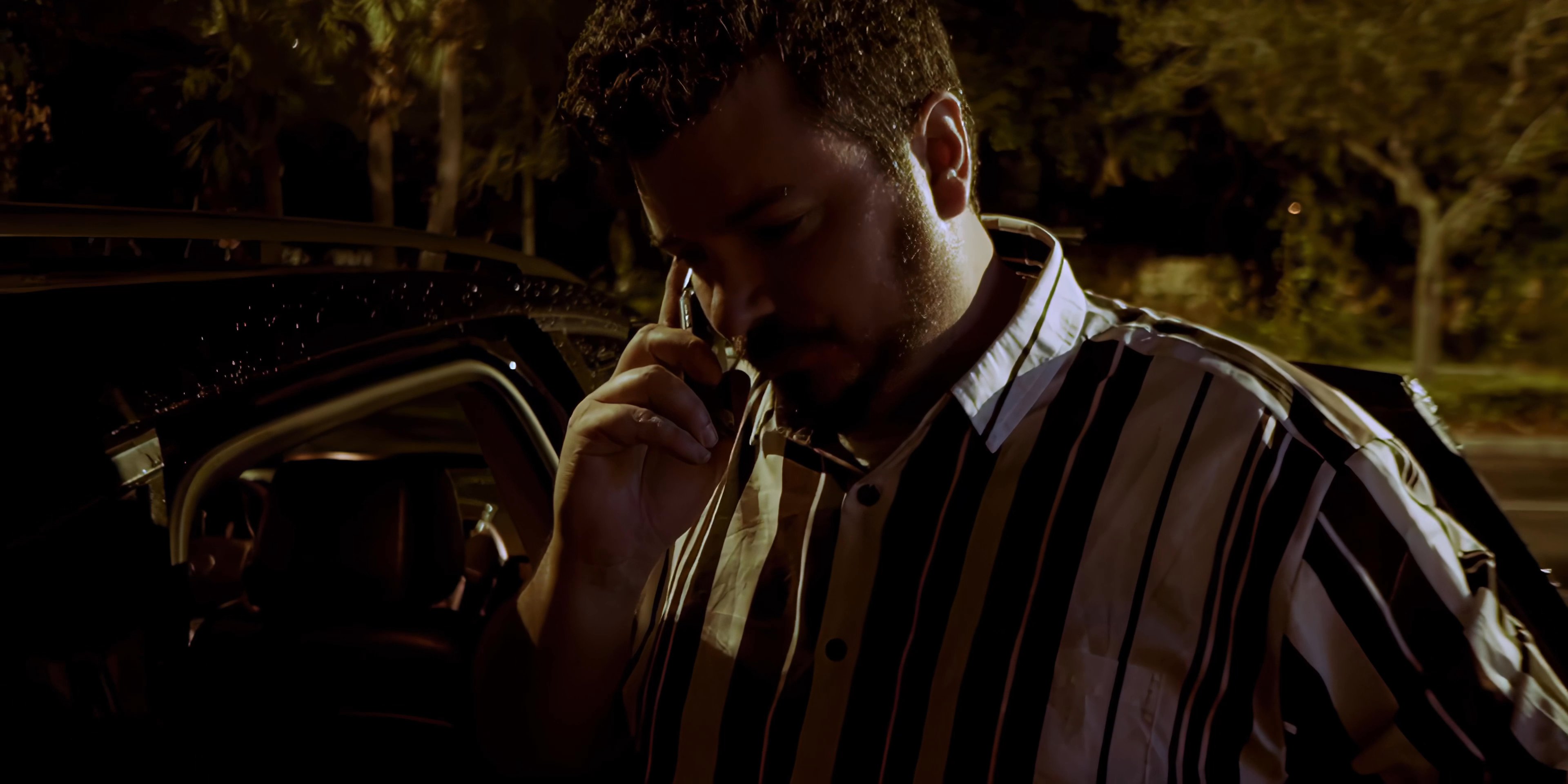 A man stands next to a car in a dimly lit parking lot during the night, engaging in a focused telephone conversation. He pauses his activity to listen intently while holding the device to his ear.