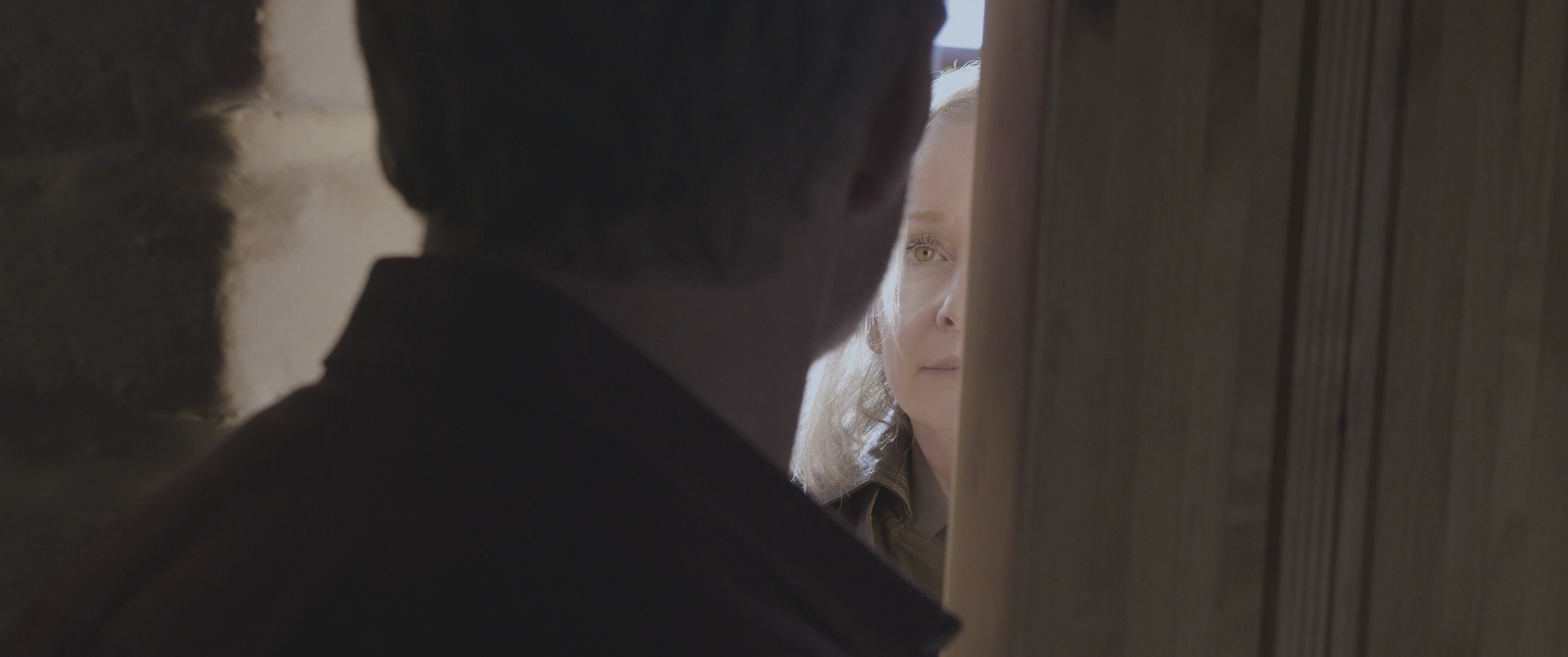 An armed individual looms in a dark doorway, creating a tense standoff with a woman on the other side. The interaction carries a clear atmosphere of threat and imminent confrontation.