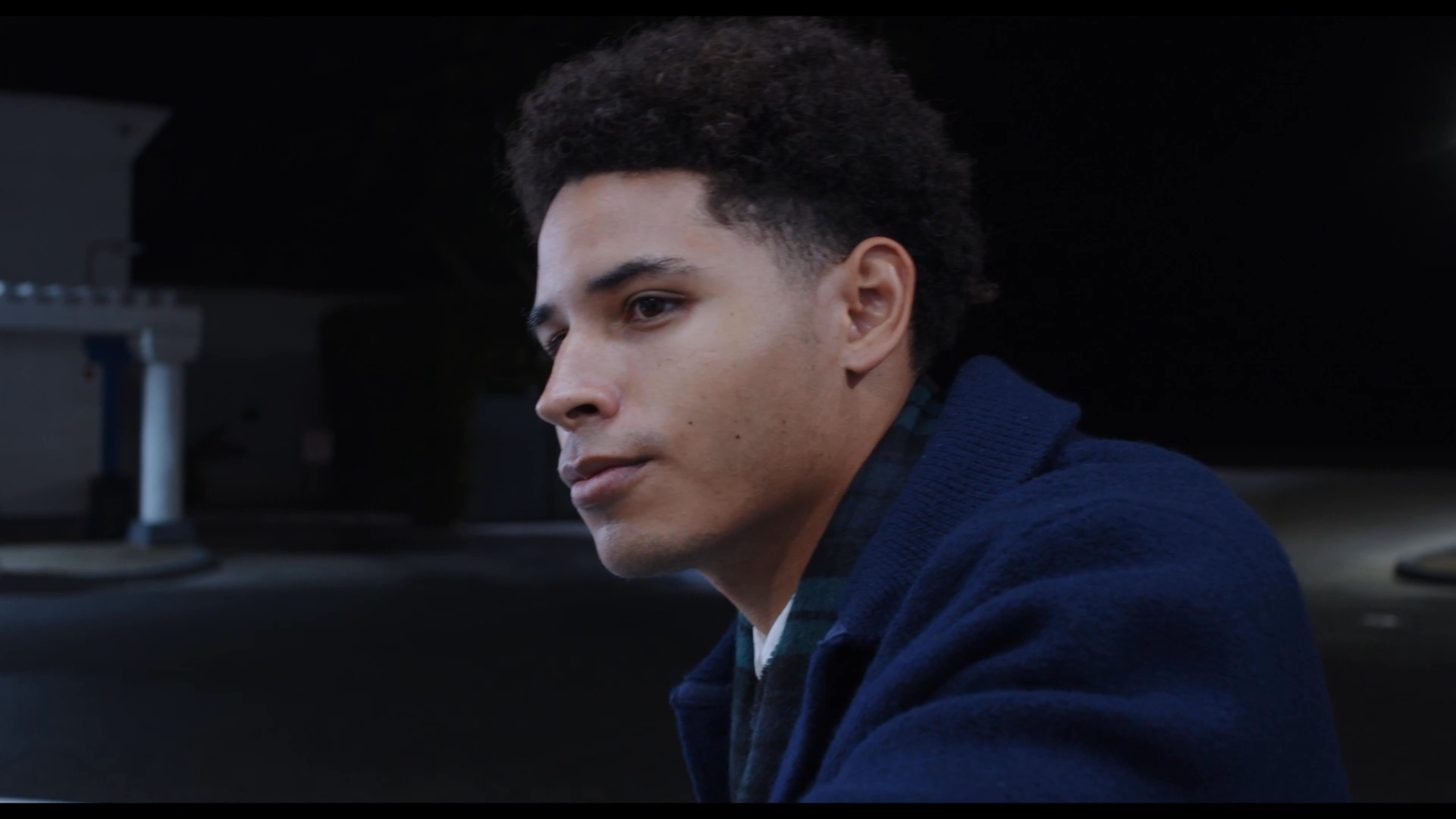 Two young men engage in conversation while standing beside a parked vehicle at a gas station during the evening hours. They share a box of breadsticks while interacting in a quiet, illuminated outdoor setting.