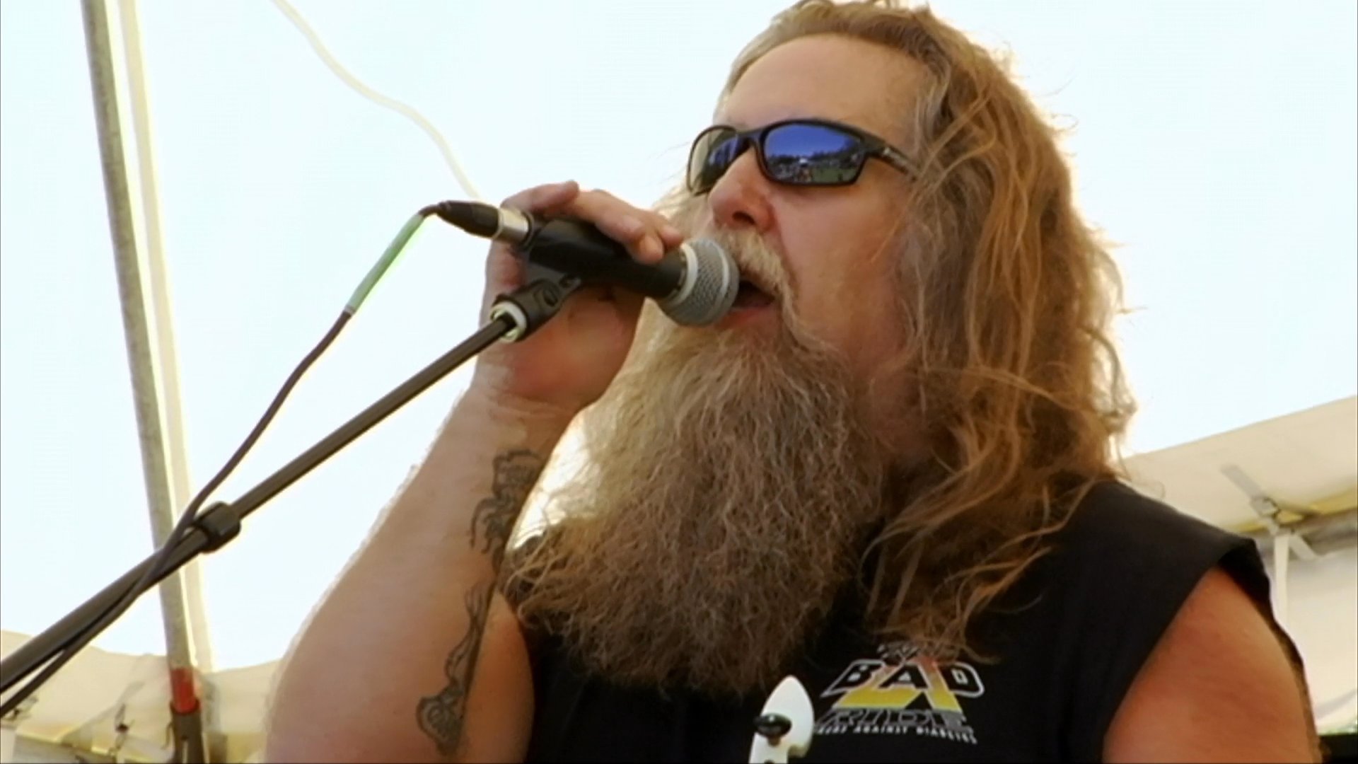 A long-haired bearded musician performs live on an outdoor stage at a biker event featuring a "Jesus Loves Bikers Too" sign. The scene captures the intersection of motorcycle culture and live music festivities.