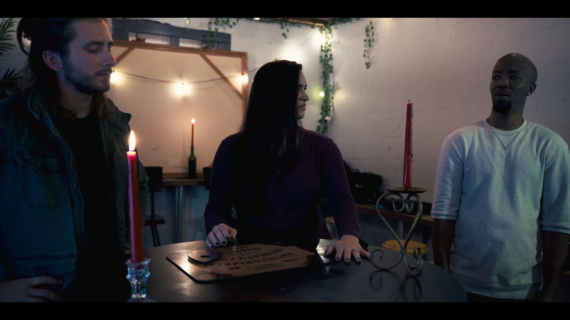 Three individuals participate in a seance, gathering around a ouija board illuminated by candlelight to communicate with spirits.