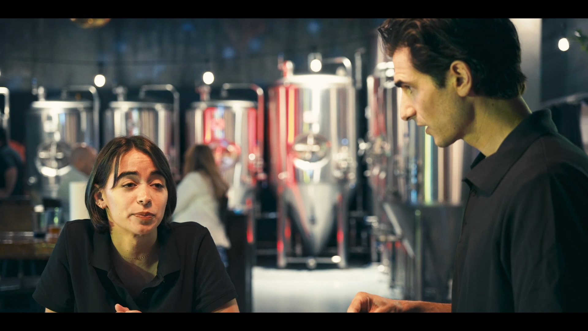 Two brewery employees engage in a focused conversation while standing at a stainless steel counter in front of large industrial fermentation tanks.