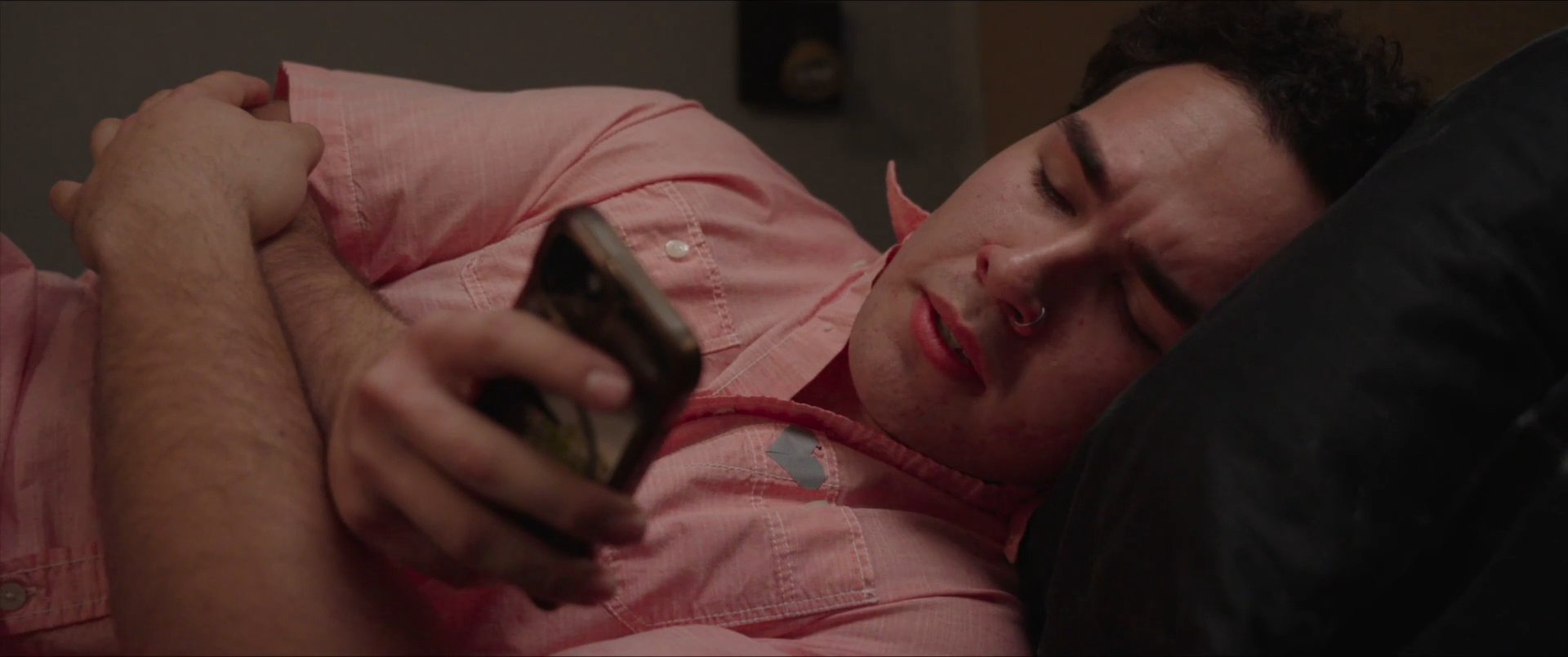 Two parents share a somber conversation in a living room while their son reclines on a couch, isolated as he scrolls through his smartphone. The contrasting activities highlight a disconnect in family communication during a period of emotional strain.