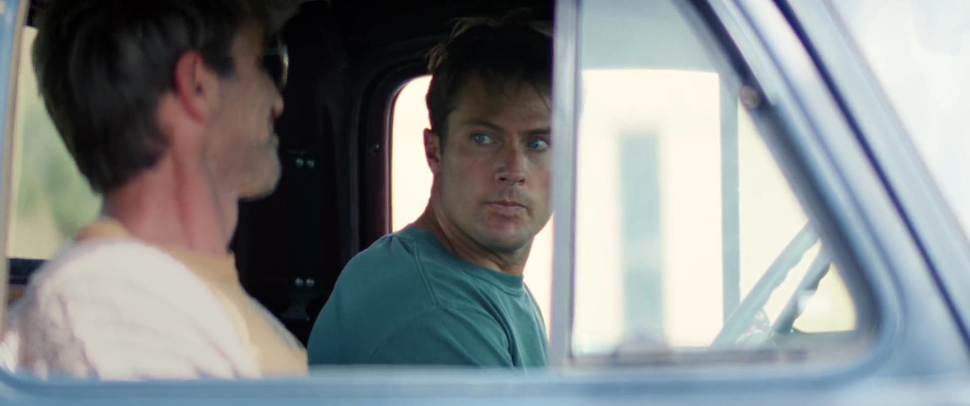 Two men sit inside a vintage truck, engaging in a relaxed and friendly conversation. The scene focuses on their interpersonal dynamic during this informal encounter.