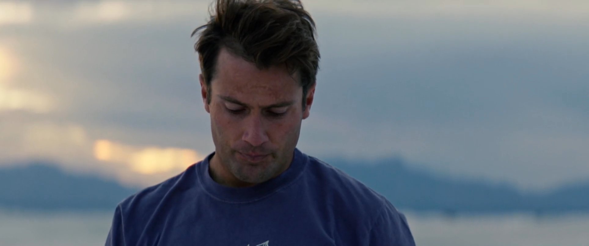 A man sits alone on white sand dunes during a sunset, reflecting on personal items left in the sand before standing to gaze at the horizon. The sequence captures a contemplative moment set against a vast, serene desert landscape.