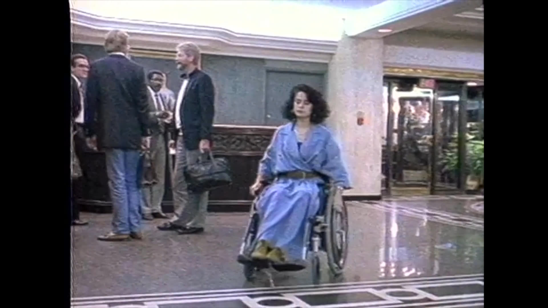 Two women converse in a monochrome setting before the scene shifts to color, where a woman in a wheelchair approaches a hotel reception desk to speak with a staff member.