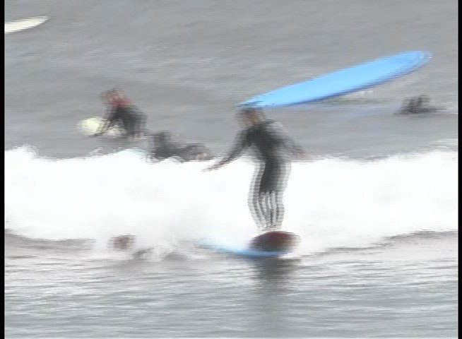 A surfer in a wetsuit maneuvers a surfboard across breaking ocean waves while other surfers paddle nearby.