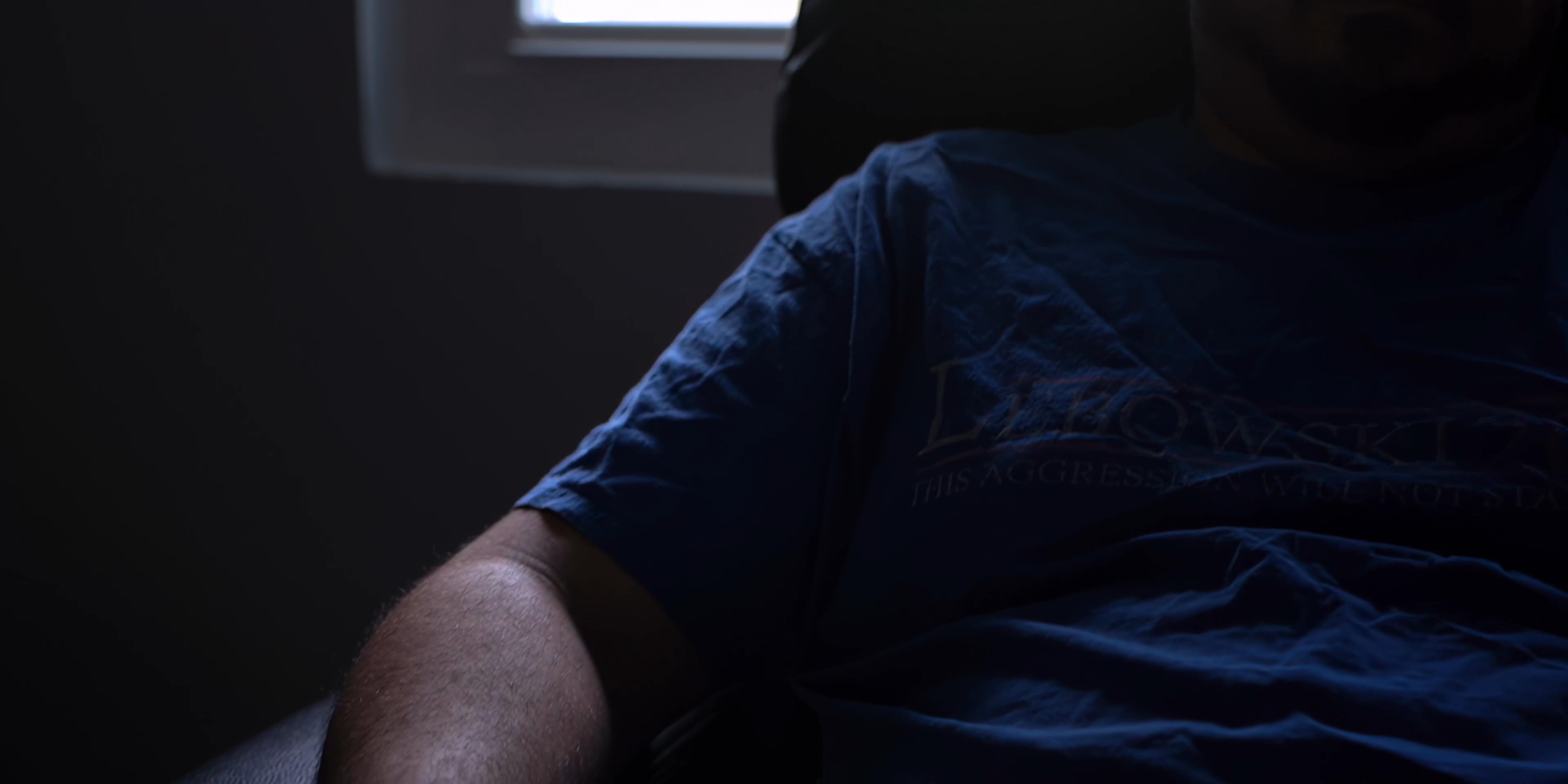 A man sits calmly in a recliner chair, holding a beverage while resting in a dim, quiet room.