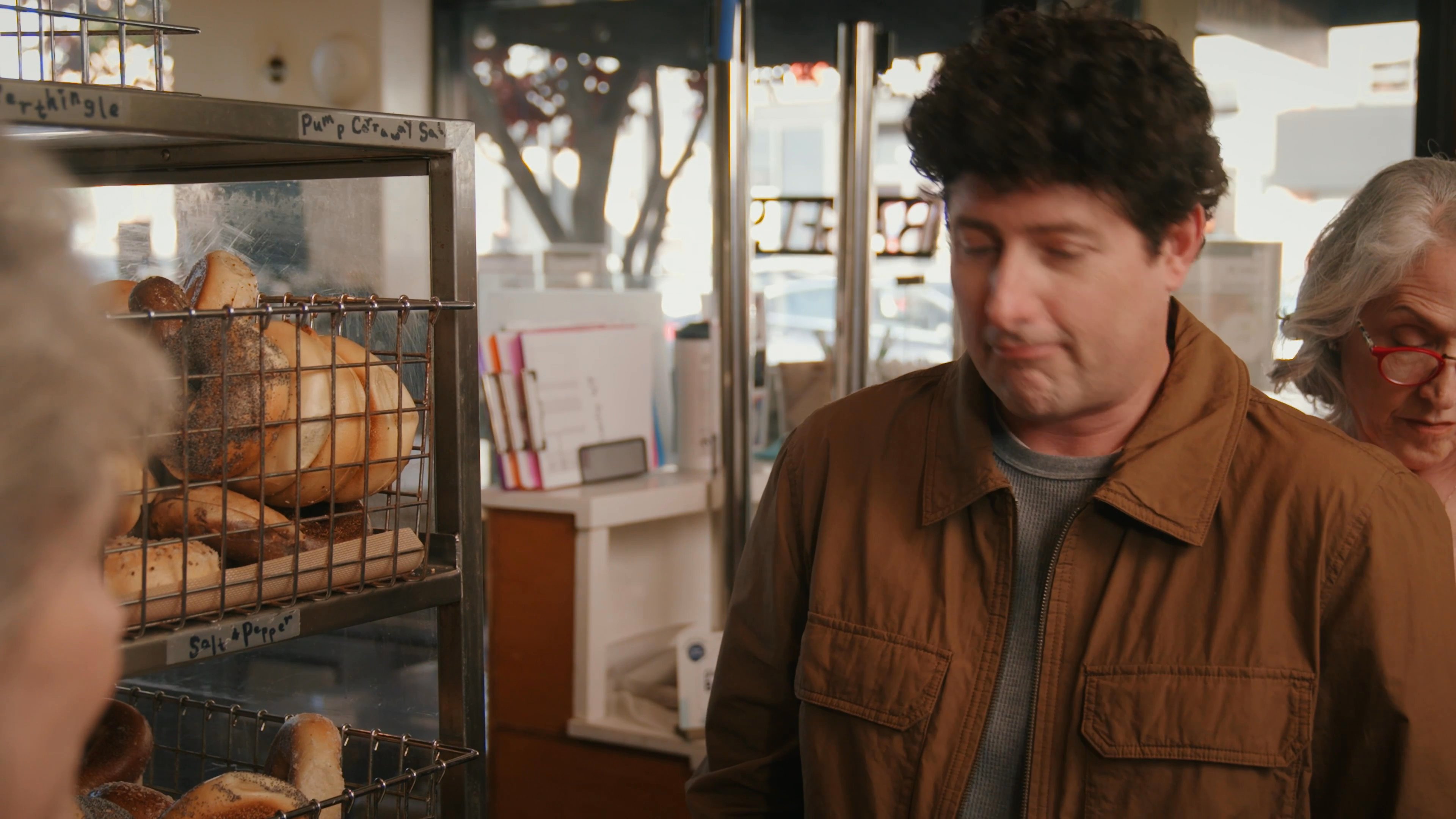 An elderly woman and a man engage in a polite, friendly conversation while standing in front of a wire rack filled with fresh bagels at a bakery.