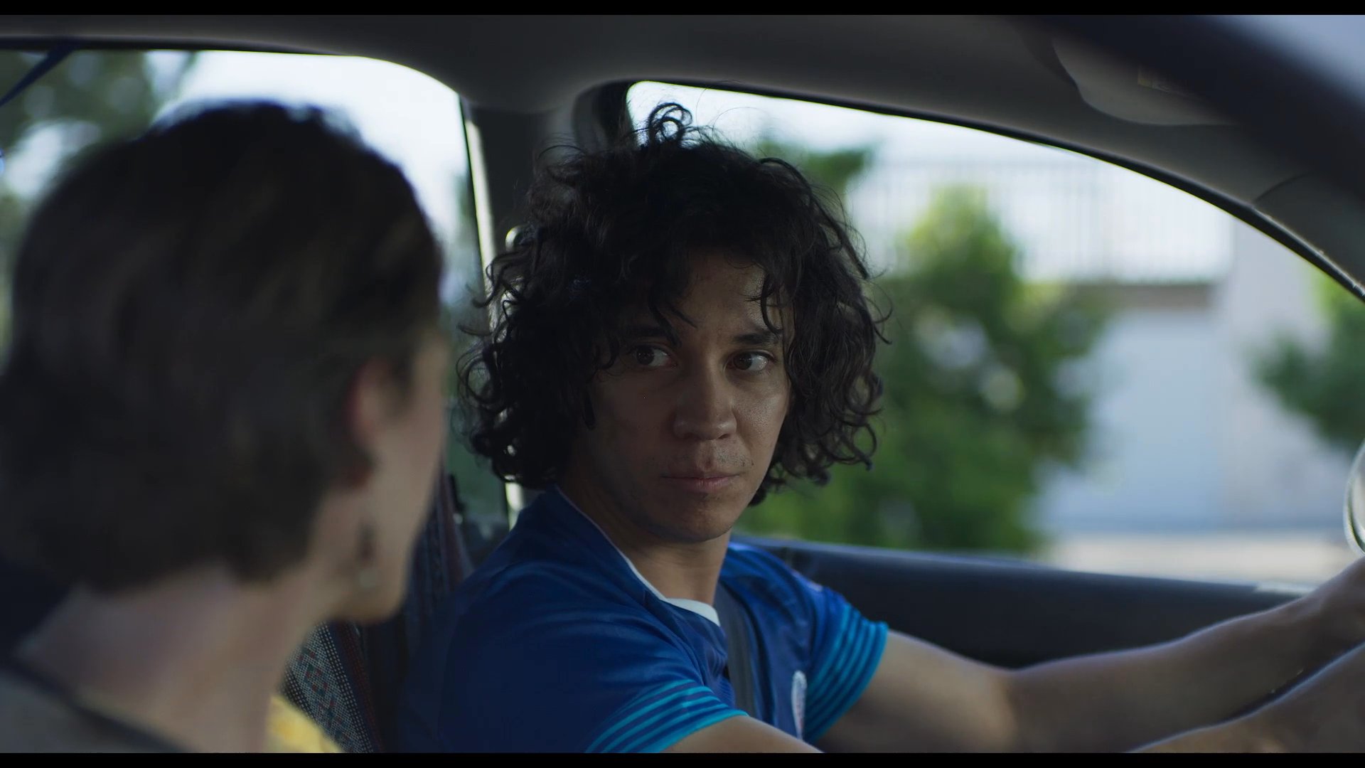 An instructor provides guidance to a young student driver while seated together inside a car. The pair discusses driving techniques before the vehicle departs down a suburban street.
