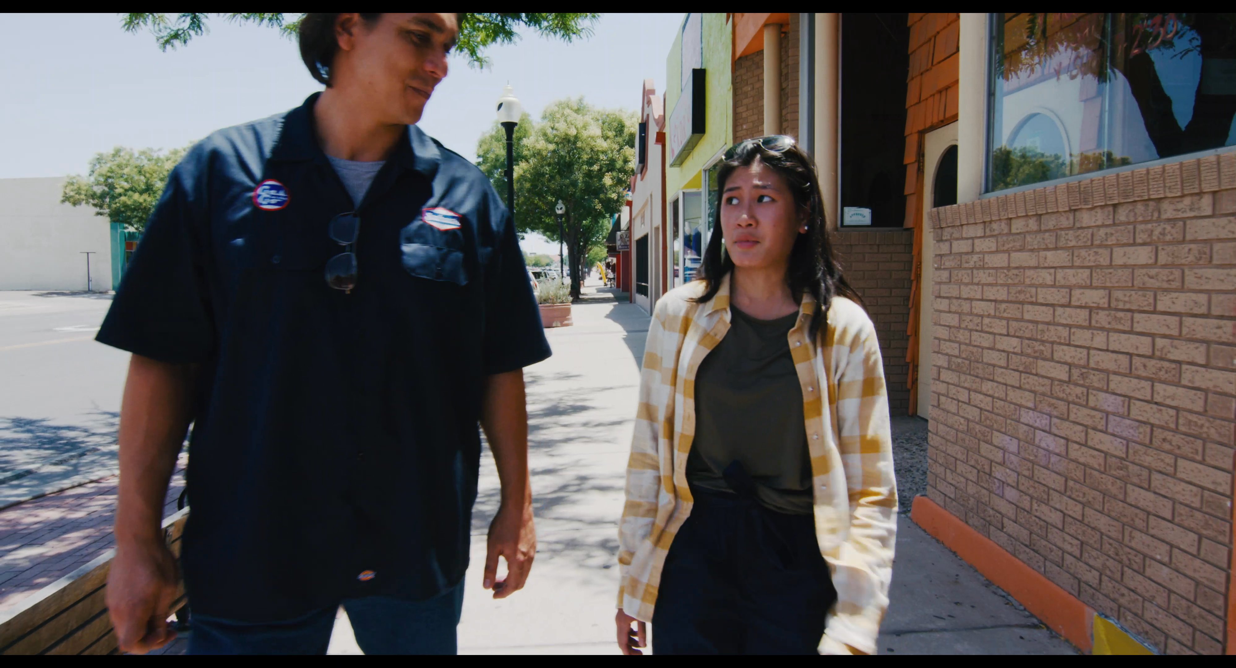 Two young adults engage in conversation while walking along a sunny urban sidewalk lined with storefronts. The pair moves at a steady pace through the outdoor pedestrian area.