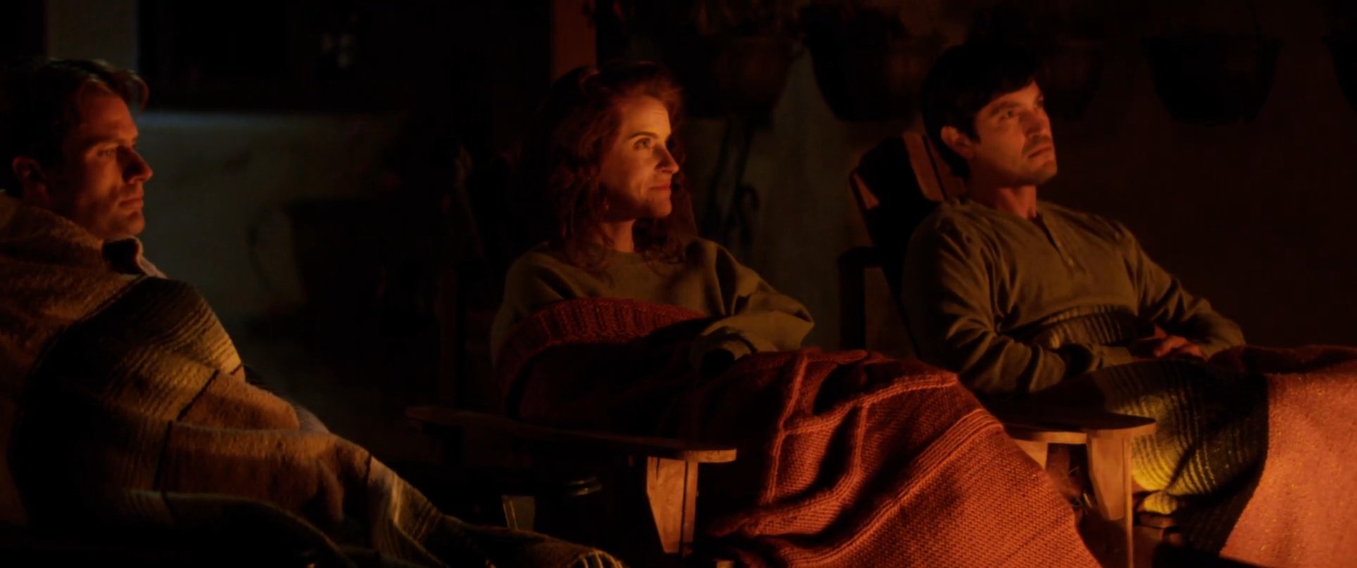 A man shares a story by a campfire while a small group listens attentively in the warm evening glow. The intimate setting highlights a moment of shared connection and quiet reflection.
