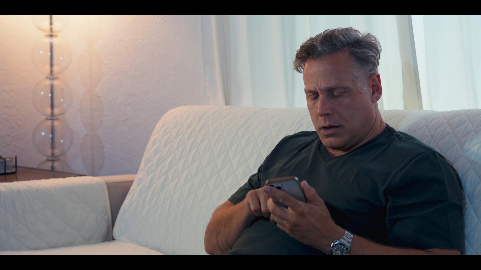 A man engages in a focused telephone conversation while seated on a sofa before transitioning to an urban environment. He observes a cityscape view and proceeds toward a building entrance marked with signage.
