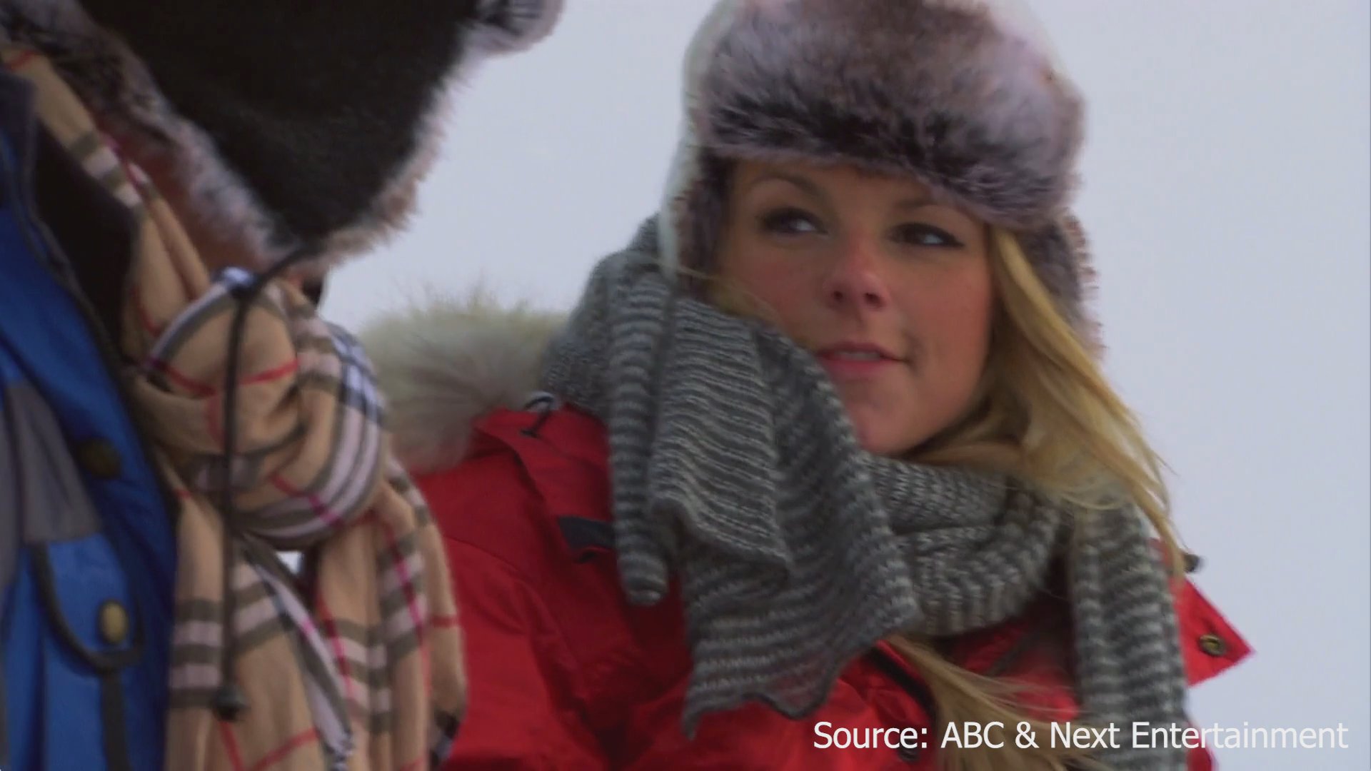 A man shows his shield and rose tattoo to a woman during an outdoor winter date in a snowy setting.