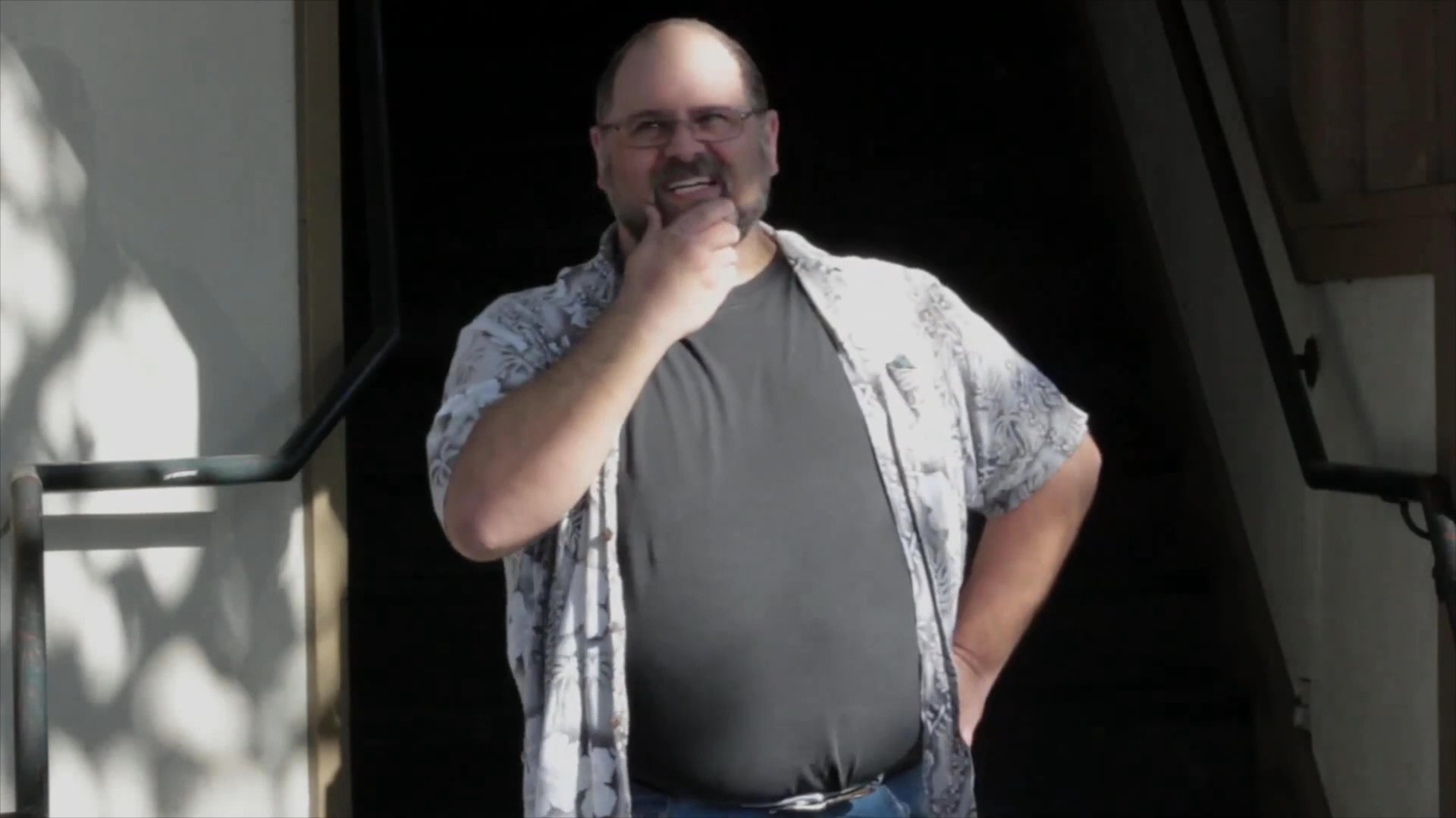 A man in a casual shirt and t-shirt stands outside a building, actively speaking and using hand gestures. He exhibits expressive body language while engaging in conversation.