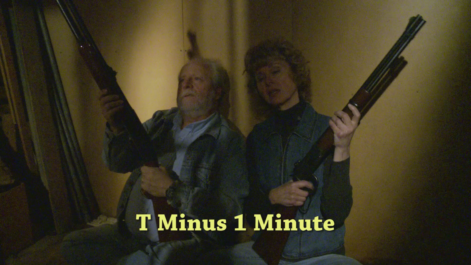 An armed couple waits in a dimly lit room, holding rifles as they countdown the final minute before a scheduled event.