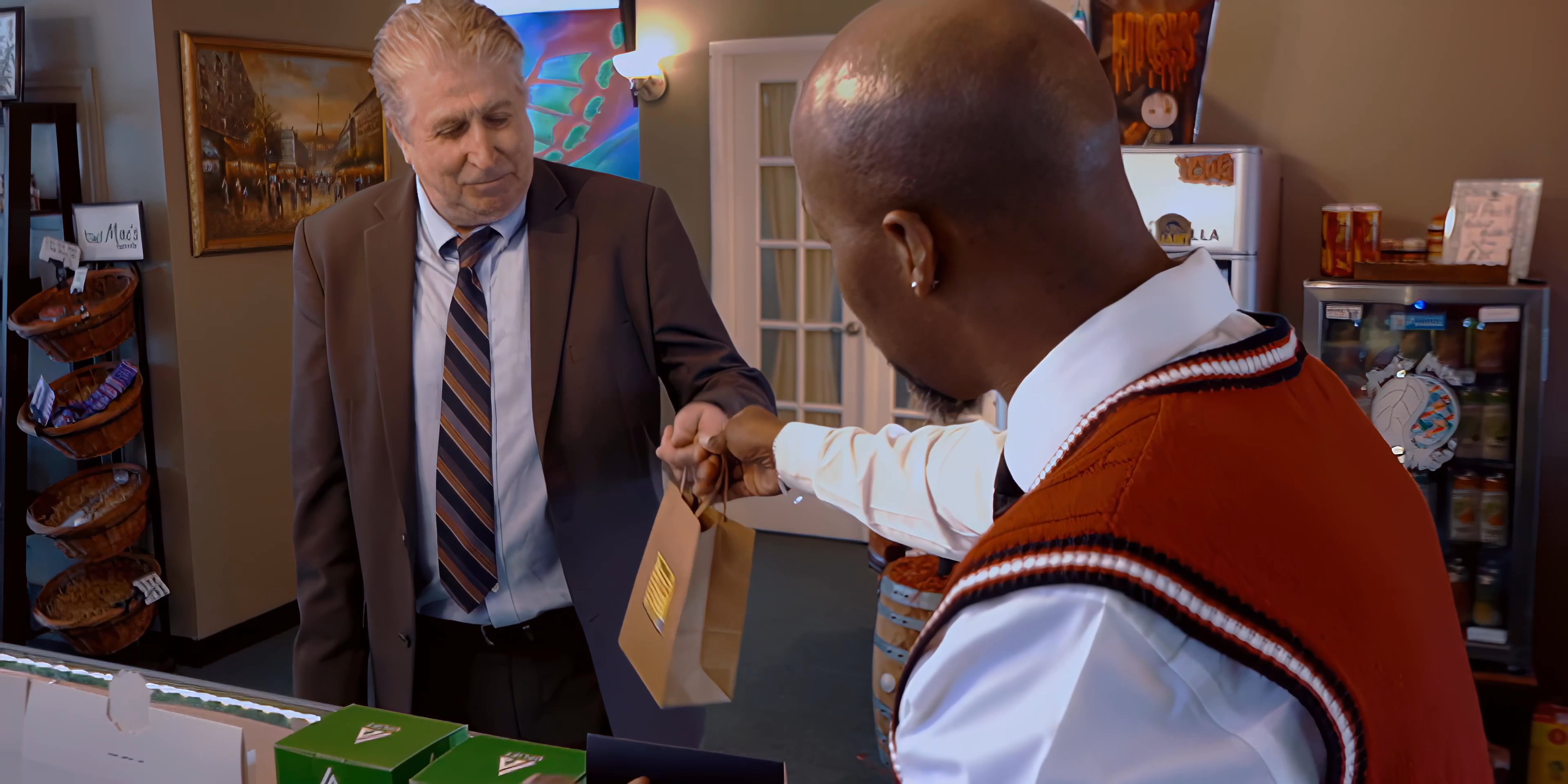 A customer in a suit completes a purchase at a convenience store counter, interacting with a clerk who retrieves and hands over a small paper bag.