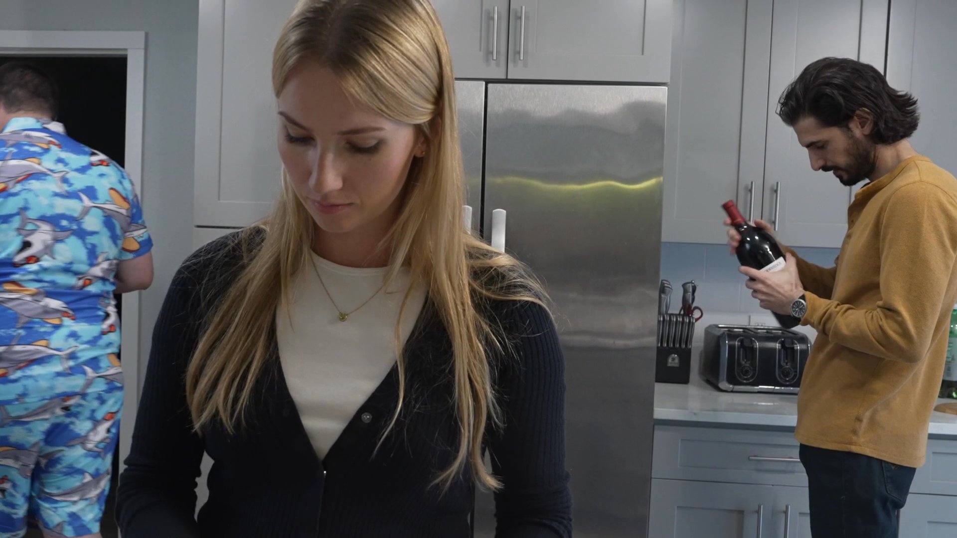 Three individuals engage in various activities within a residential kitchen, with one man examining a bottle of wine while others interact with their mobile devices and food preparation.