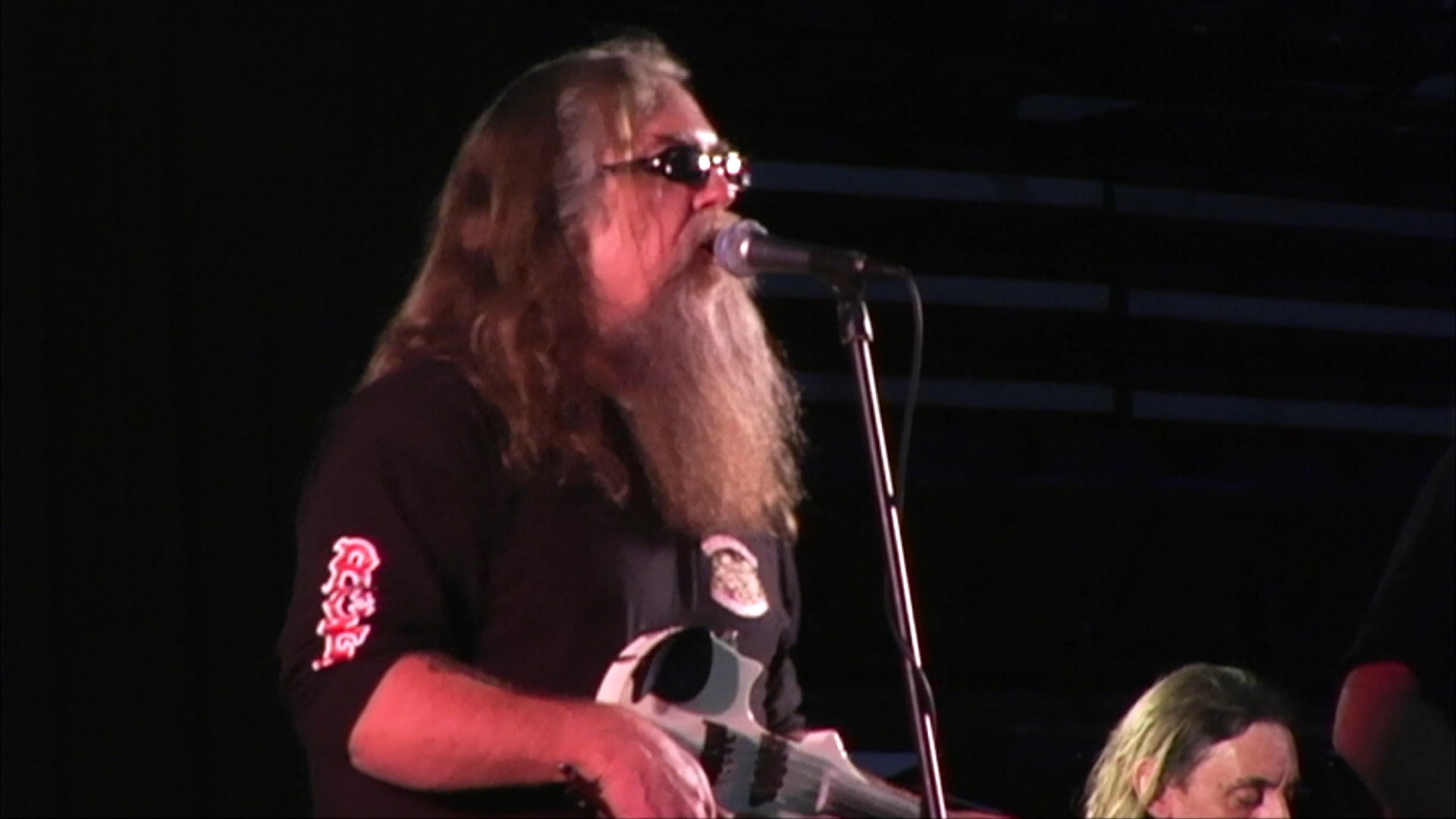 A long-haired, bearded violinist sings and plays his instrument during a live musical performance on a dimly lit stage. The musician delivers an expressive vocal and instrumental piece while accompanied by a band.