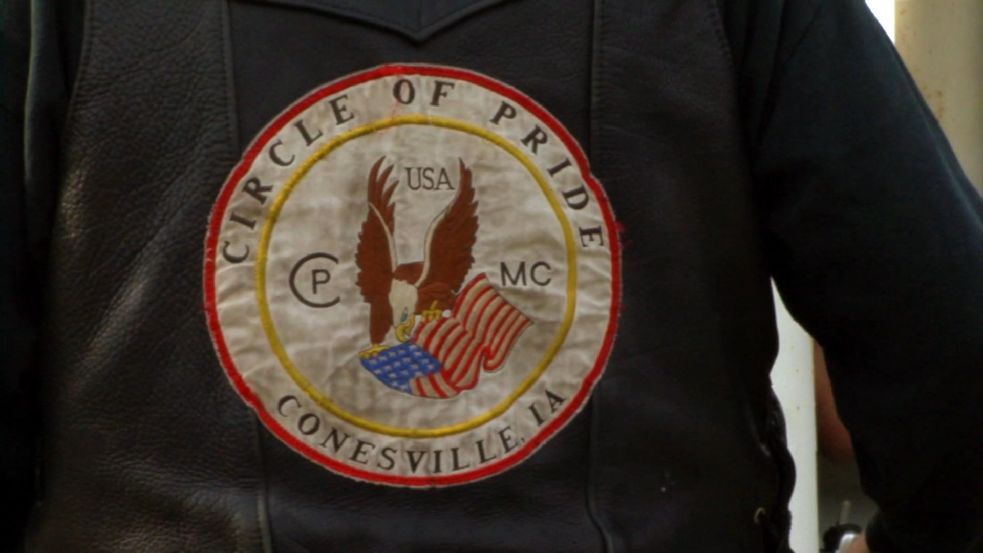 A man wearing a biker vest shares his perspective at a motorcycle rally while attendees mingle and socialize in the background.
