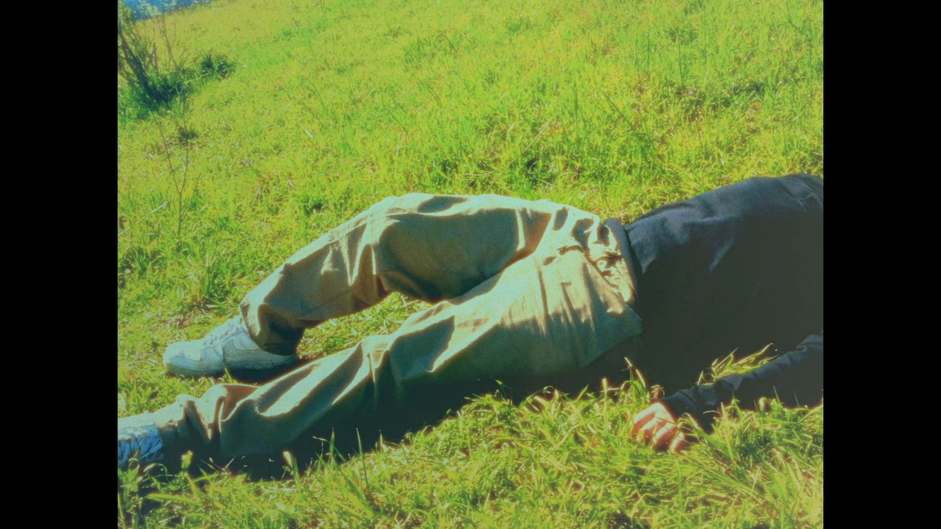 A young man finds a moment of respite while lying in a sunlit, grassy meadow. The perspective shifts from the expansive sky to a private moment of quiet reflection.