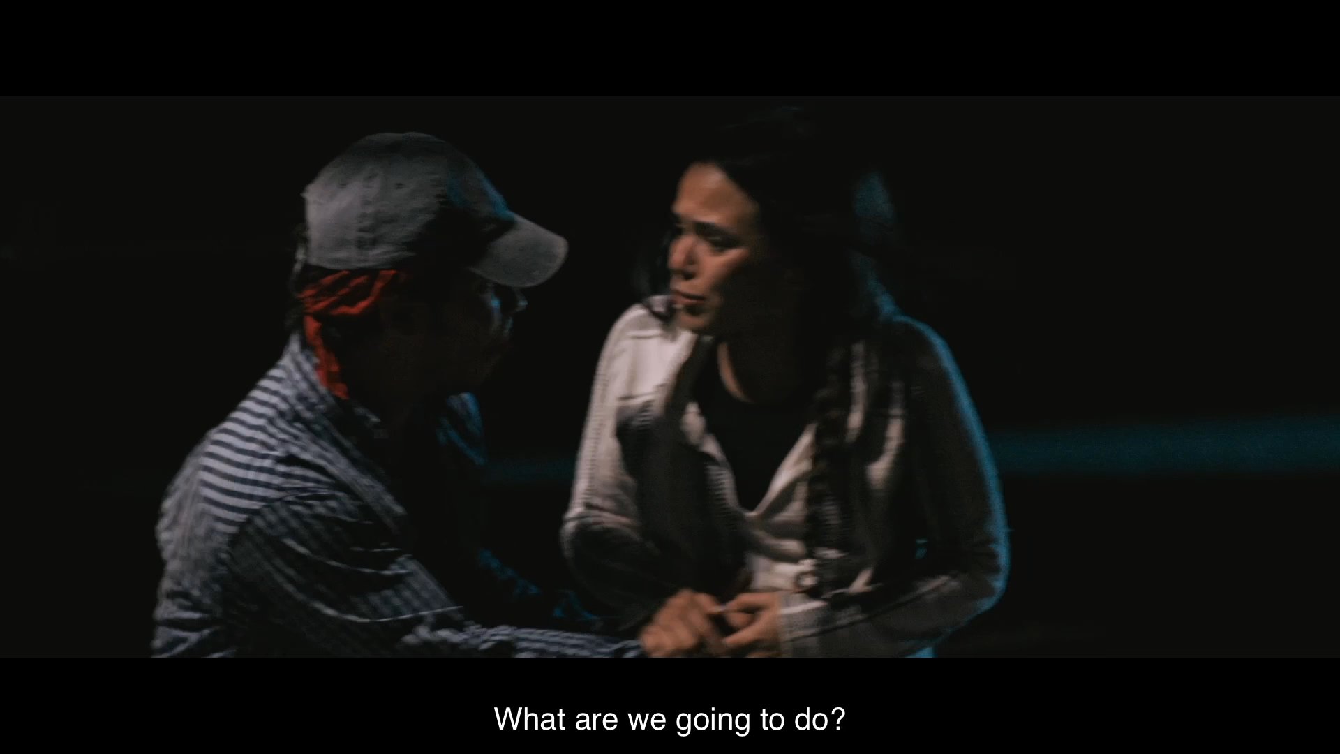 Two individuals sit together in darkness, reeling from the theft of their backpack and anxiously questioning their next move.