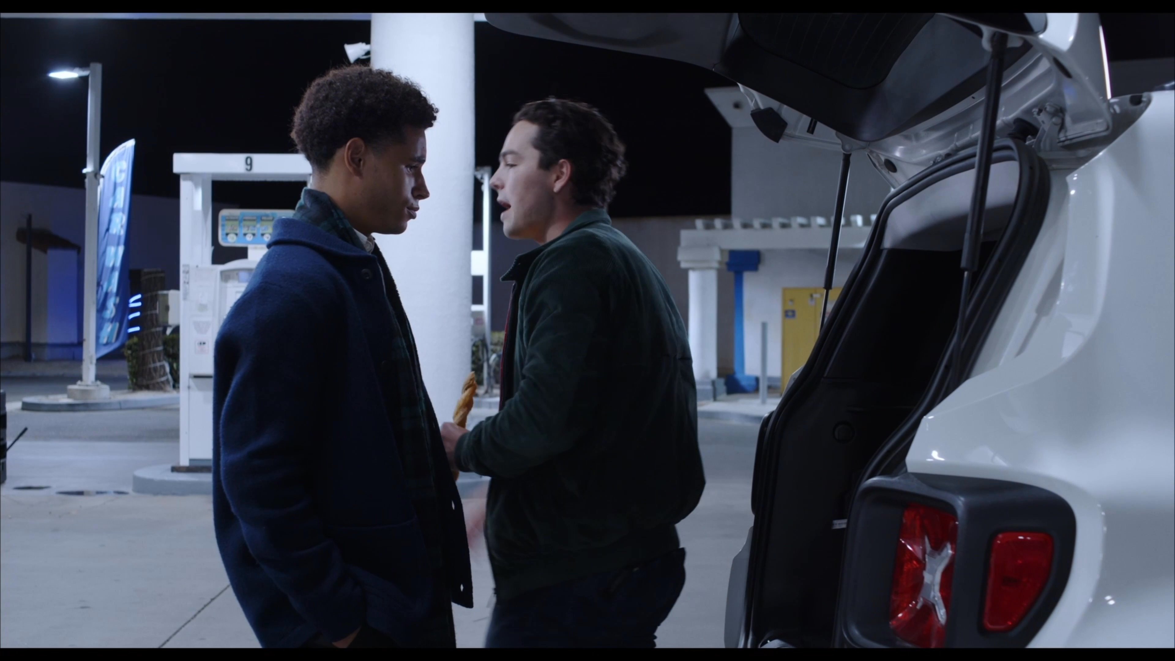 Two men engage in conversation while parked at a gas station during the night, with one individual holding a snack.