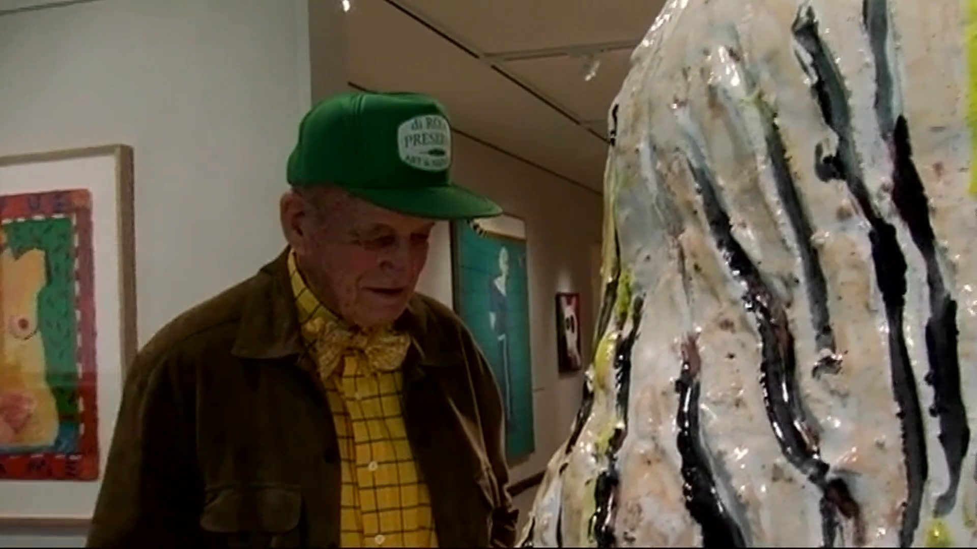 An elderly man in a green cap studies a large, textured abstract sculpture in a contemporary art gallery. Visitors browse nearby while he reflects on the artwork and its lack of an accompanying name.