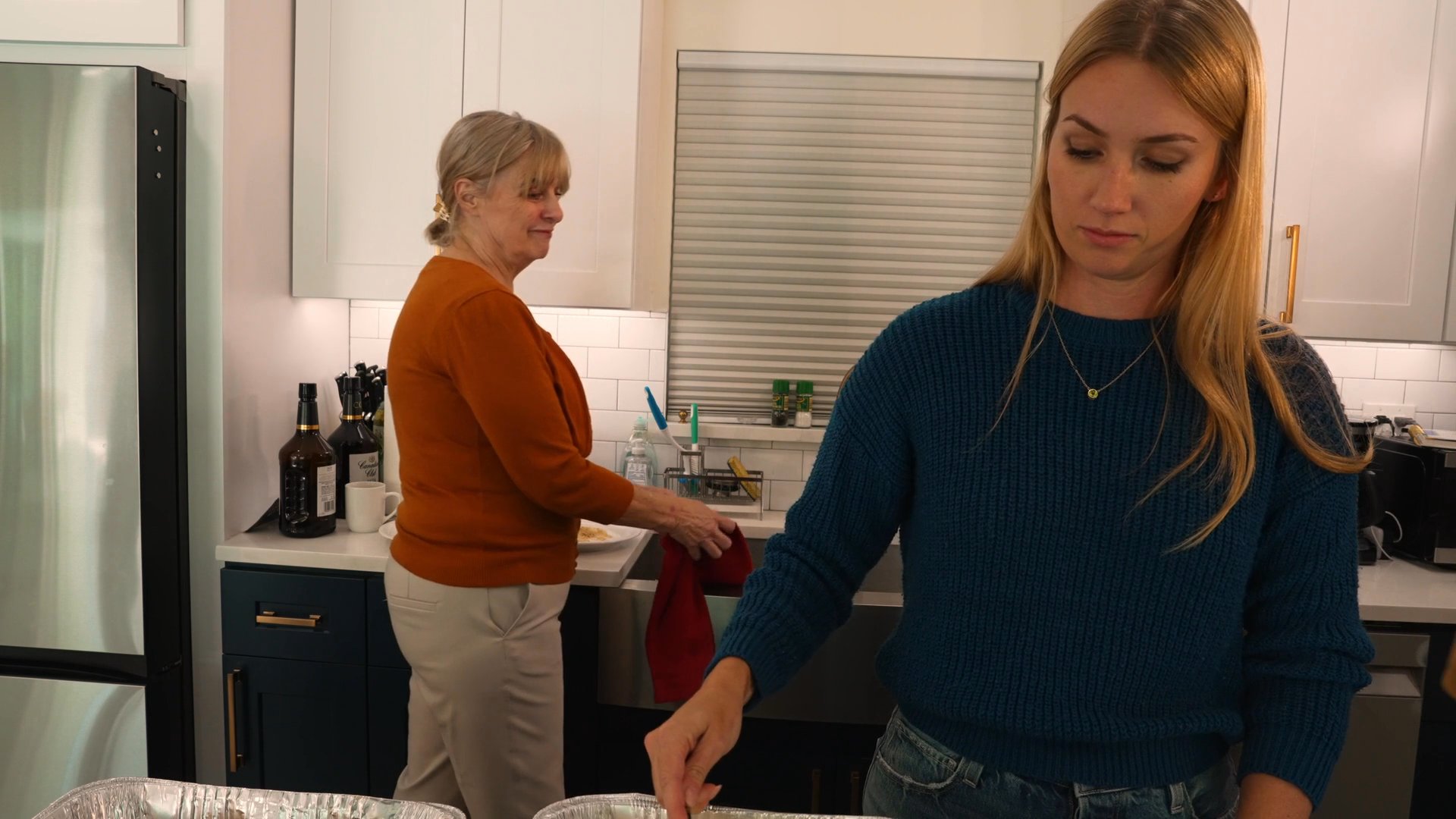 A group of people interact and prepare food within a modern kitchen setting. The scene captures a collaborative social atmosphere during a domestic gathering.
