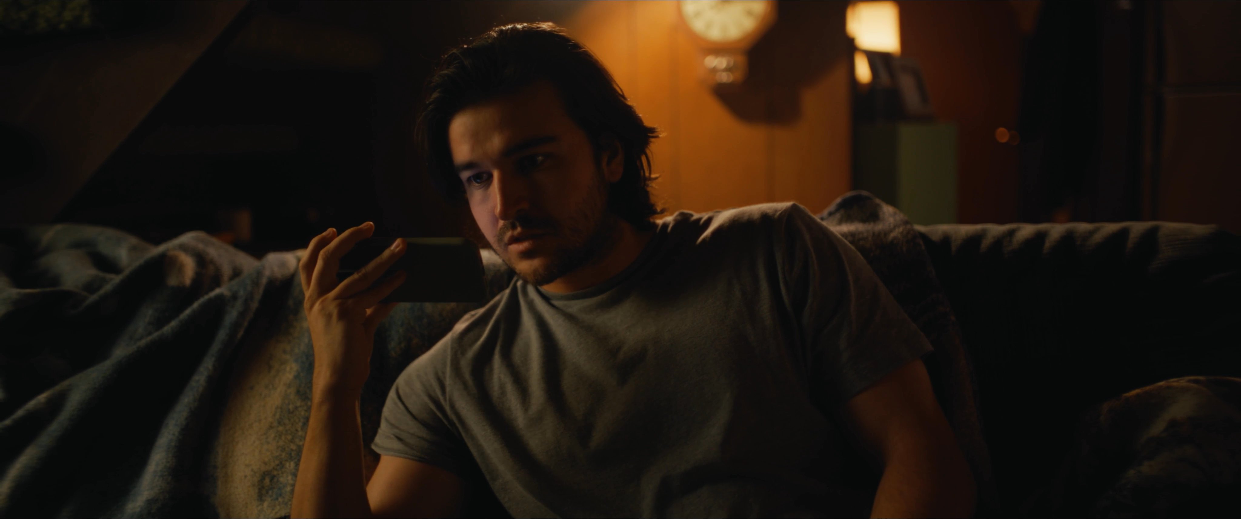 A man sits alone on a couch in a dimly lit living room, holding a bottle and browsing content on his smartphone. He appears pensive and focused as he engages with his device in the quiet setting.