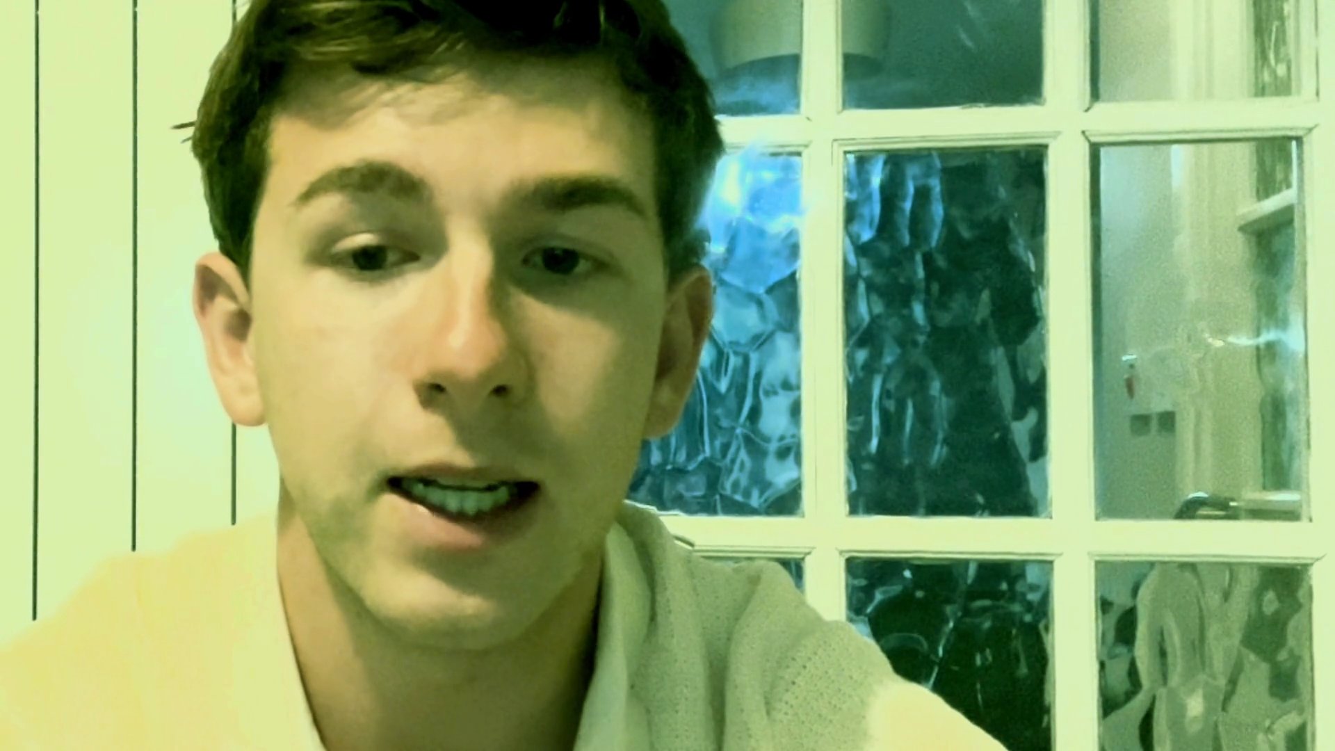 A young man speaks directly to the camera in a brightly lit indoor setting, providing commentary or a personal update.