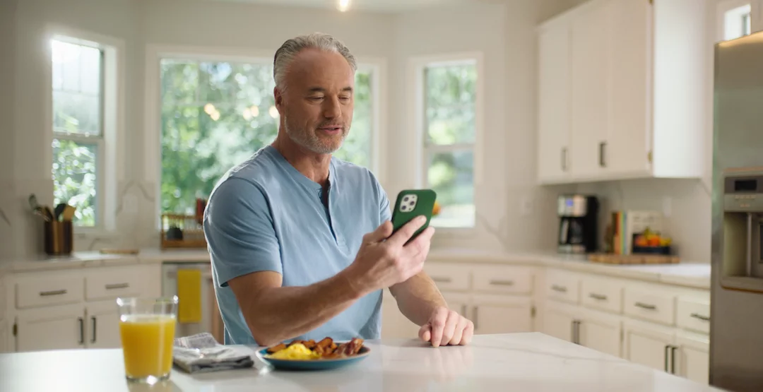 An older man sits on a couch and smiles while he looks down at his phone.