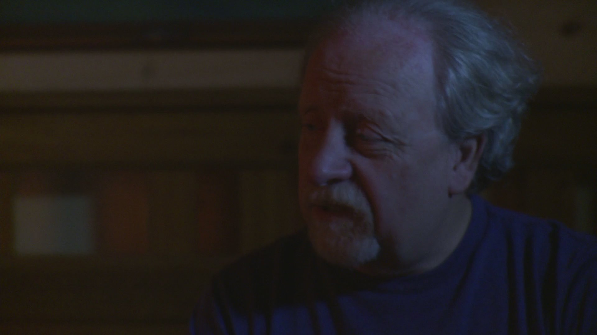A man and a woman engage in a serious, quiet conversation while sitting together on a bed in a dimly lit bedroom.