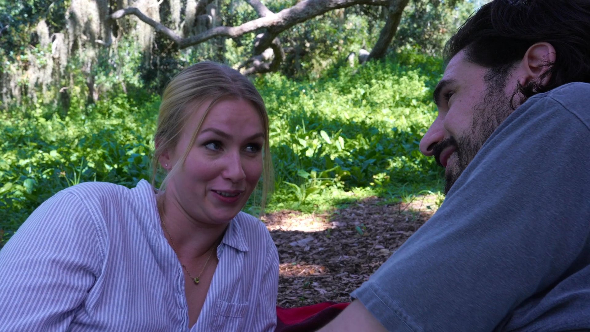 A man and woman engage in a relaxed, intimate conversation while reclining together on a blanket in a sunlit, wooded area.