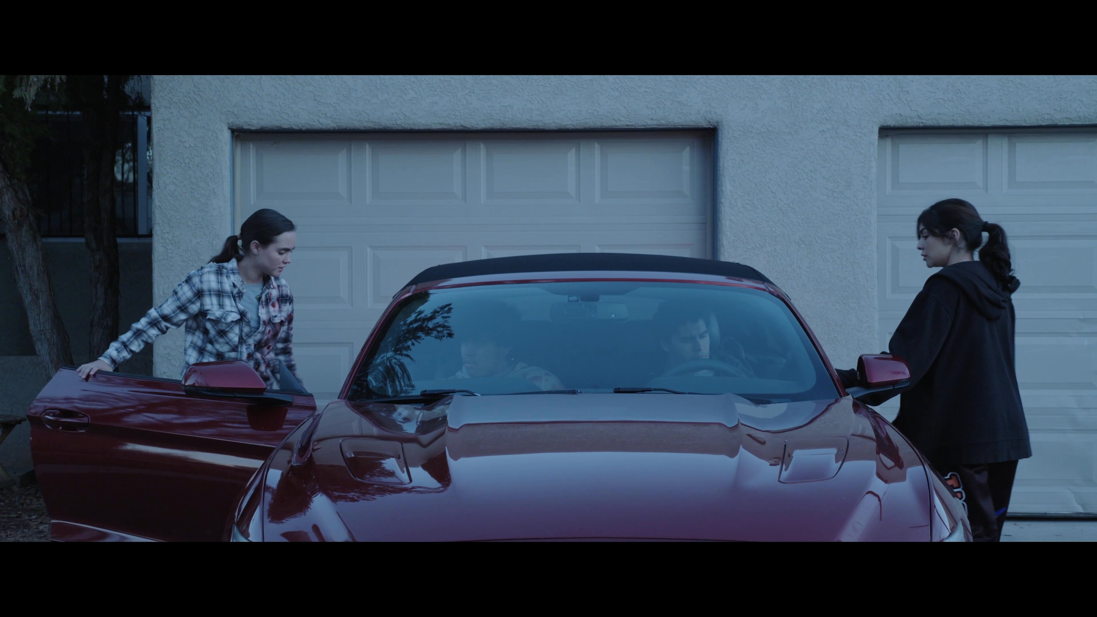 Two individuals assist a bloodied, injured young man as he exits a vehicle in front of a residential garage. The scene concludes with a tense, emotional embrace between the injured youth and his companion.
